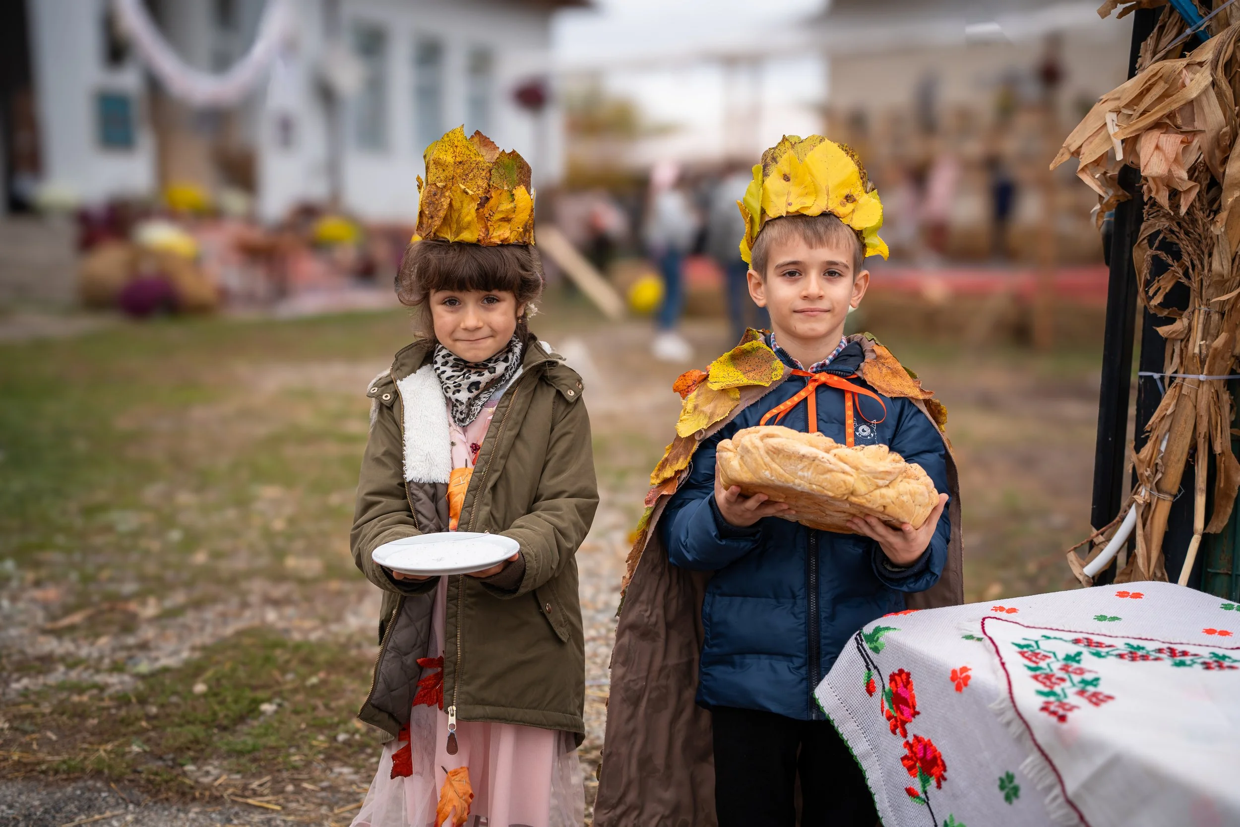 Two children standing outdoors during autumn festival, wearing crowns made of colorful leaves, holding baked goods, one with a plate and the other with a bread loaf, near a table with embroidered cloth.