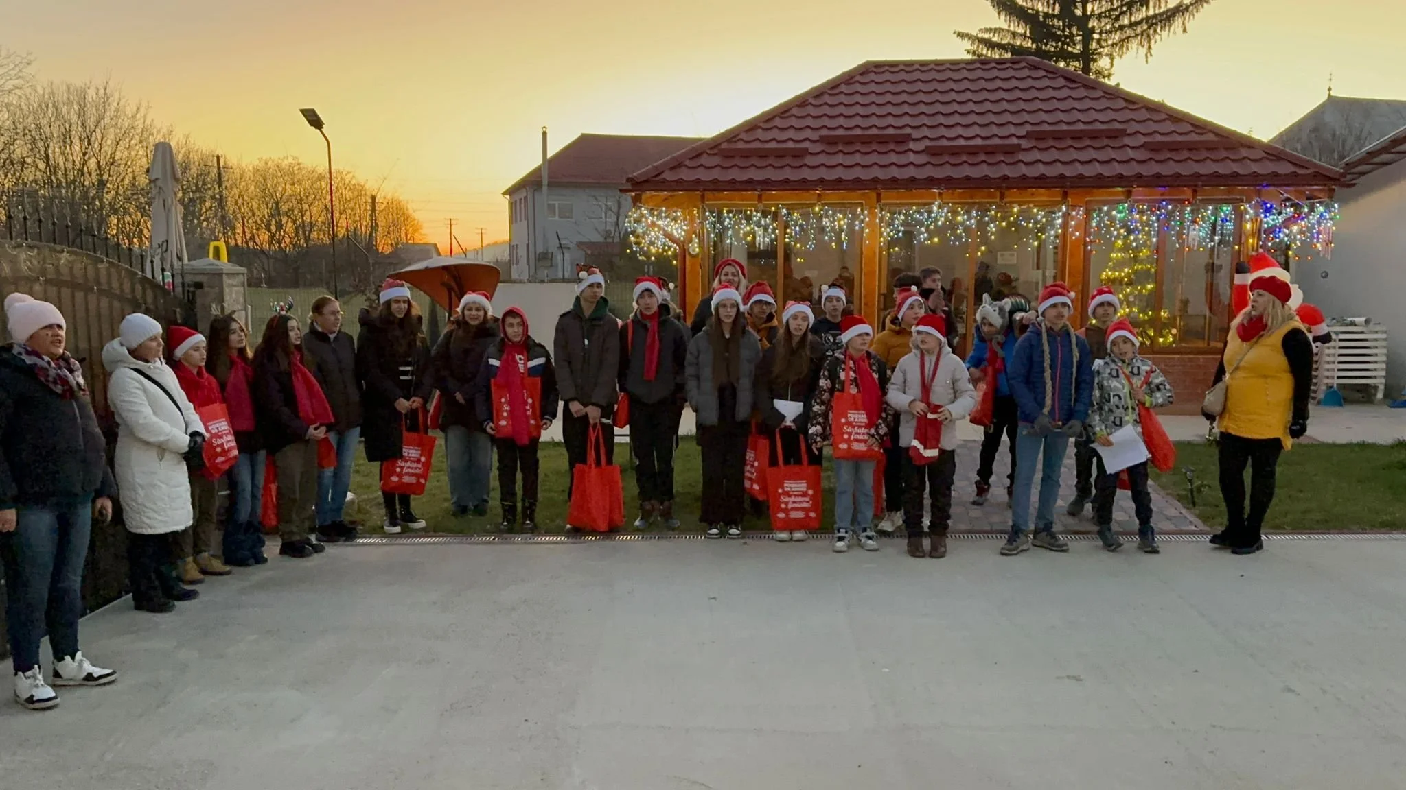 Group of children and adults in Christmas hats gathered outside a decorated wooden pavilion with string lights, during sunset.