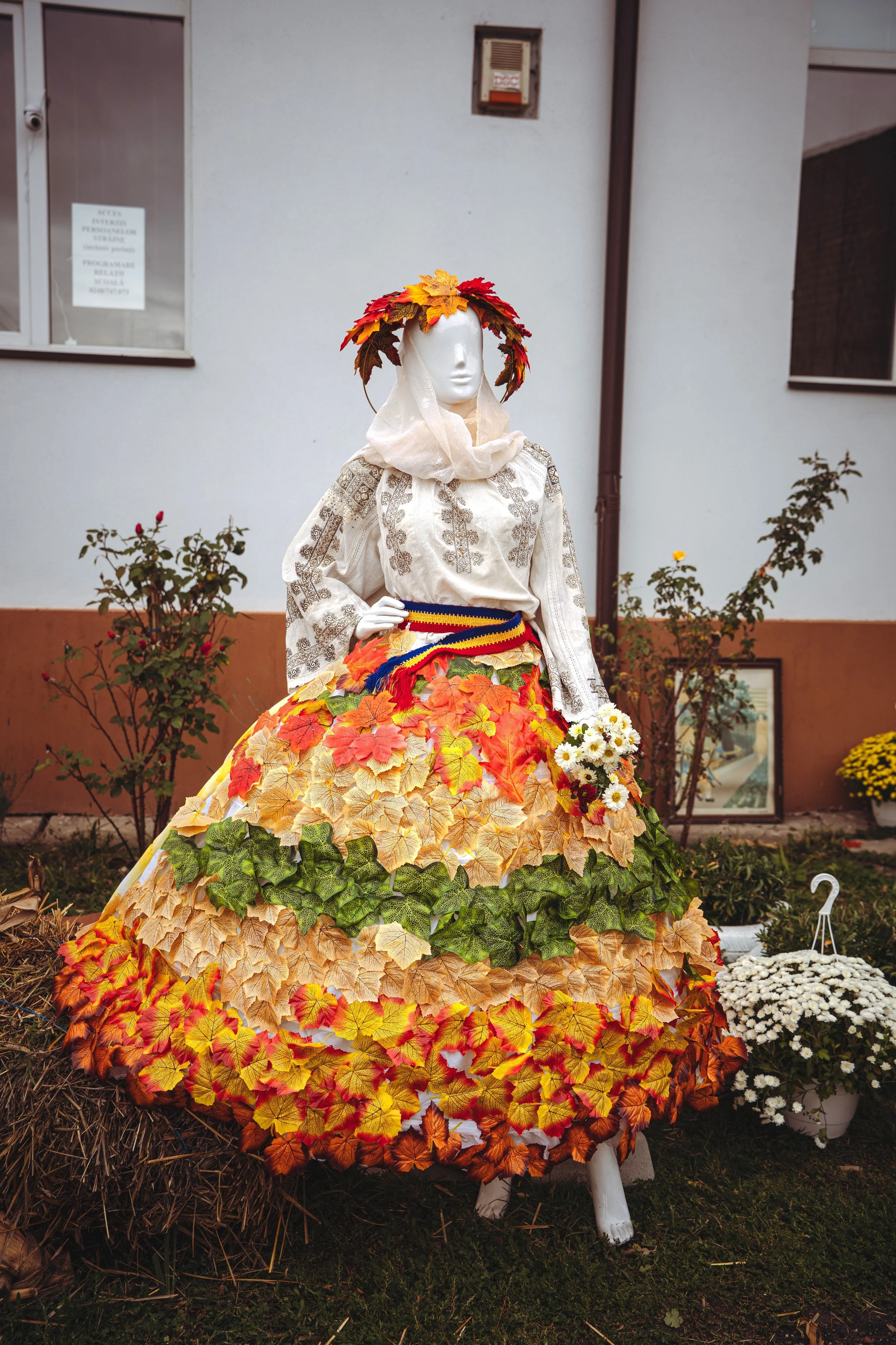 A mannequin dressed in a traditional folk outfit with a skirt decorated with artificial fall leaves in shades of orange, yellow, and green. The mannequin has a head adorned with a crown of artificial fall leaves and holds a small bouquet of white flo