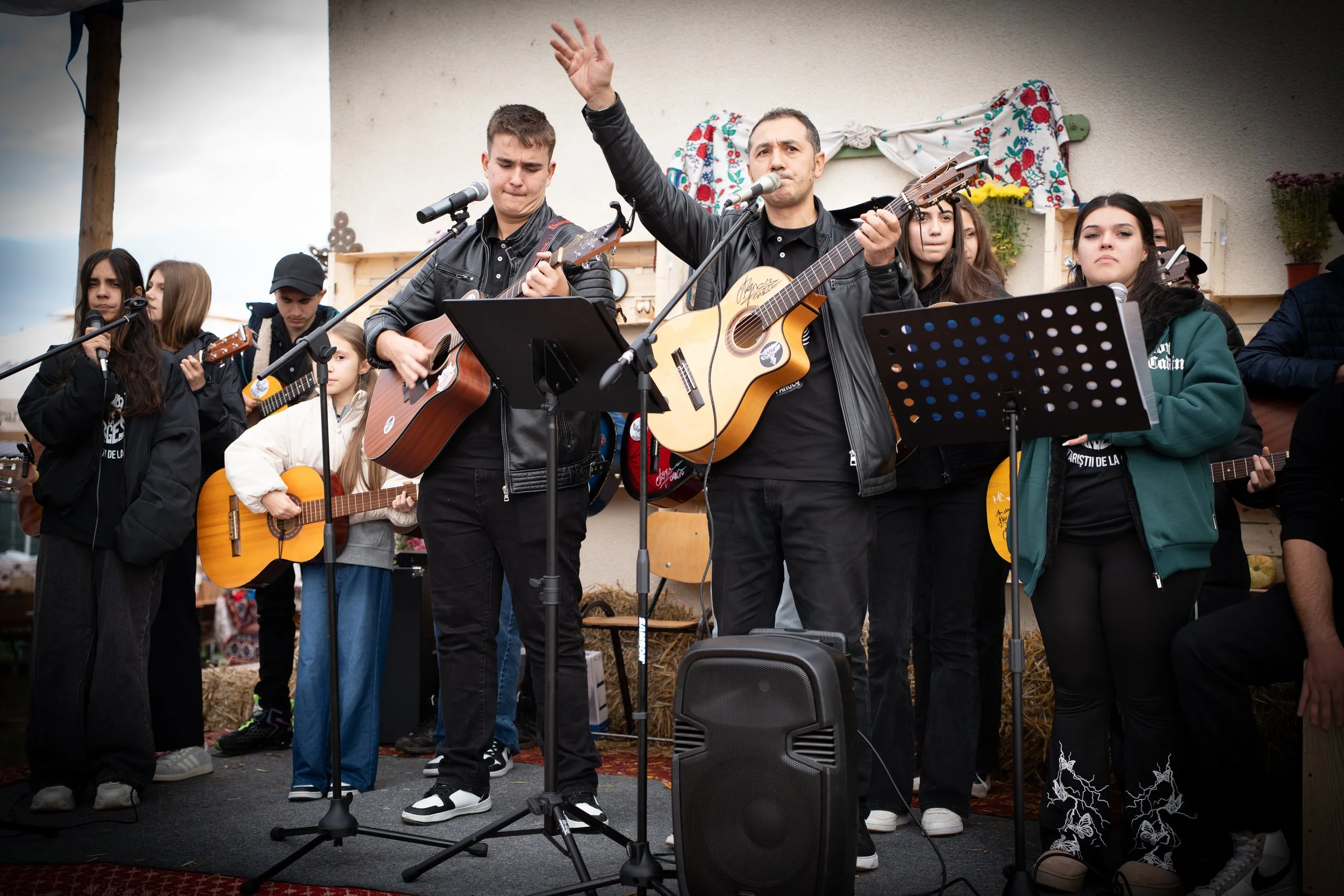 Group of people singing and playing guitars on stage, with a man in the center raising his hand, at an outdoor event.