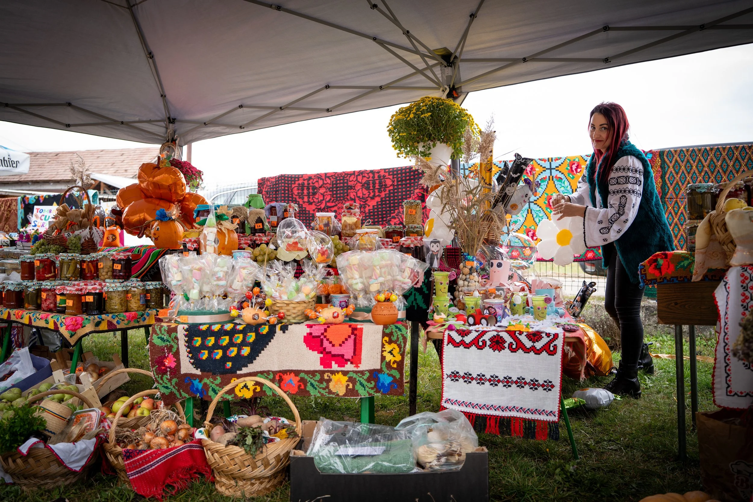A woman standing behind a market stall under a canopy, selling a variety of colorful homemade crafts and food items, including decorated pumpkins, jars, baskets of apples, and woven items, with vibrant embroidered textiles in the background.