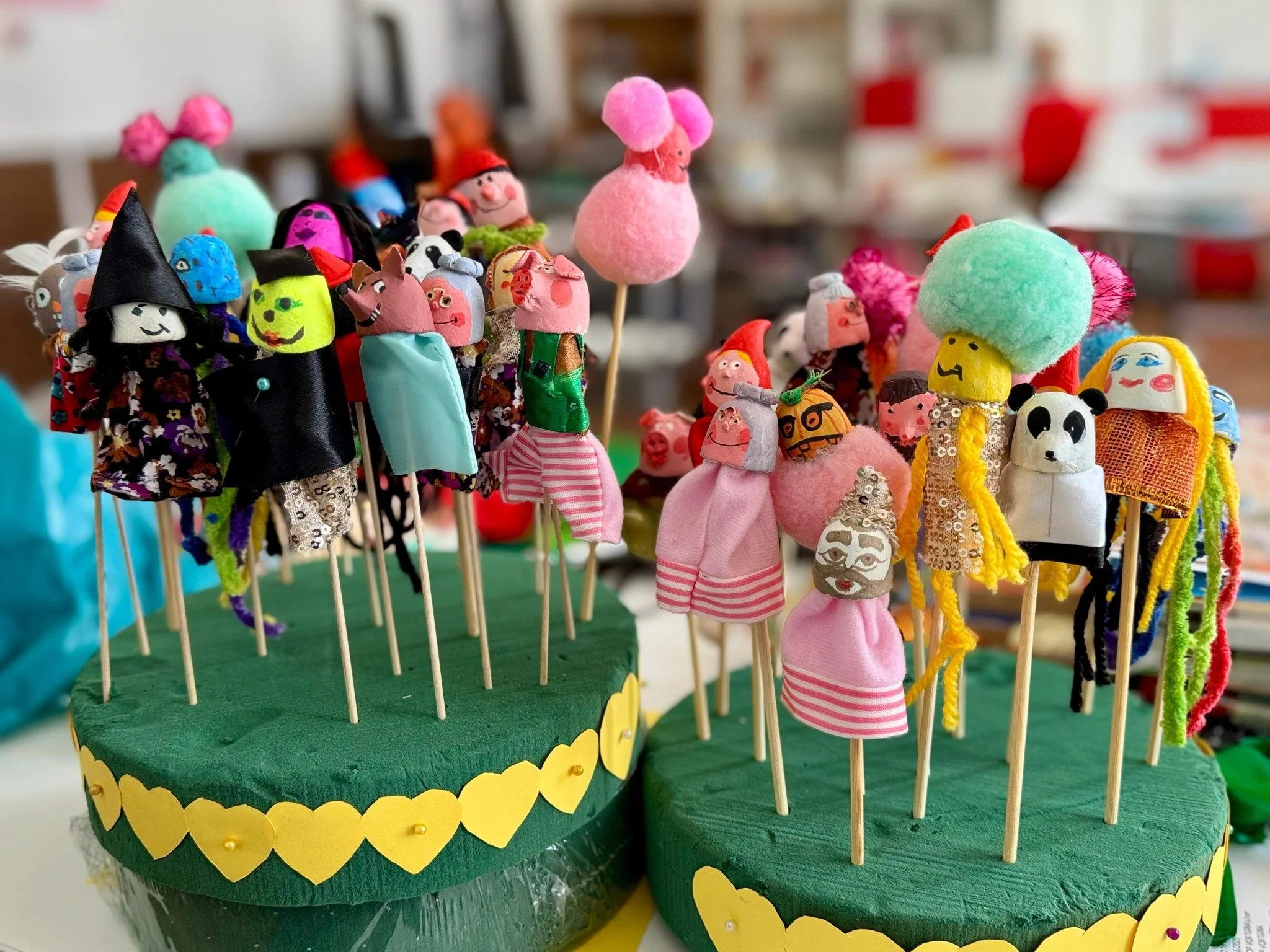 Colorful puppet crafts on sticks resembling various characters, displayed on two green foam bases decorated with yellow heart-shaped cutouts.