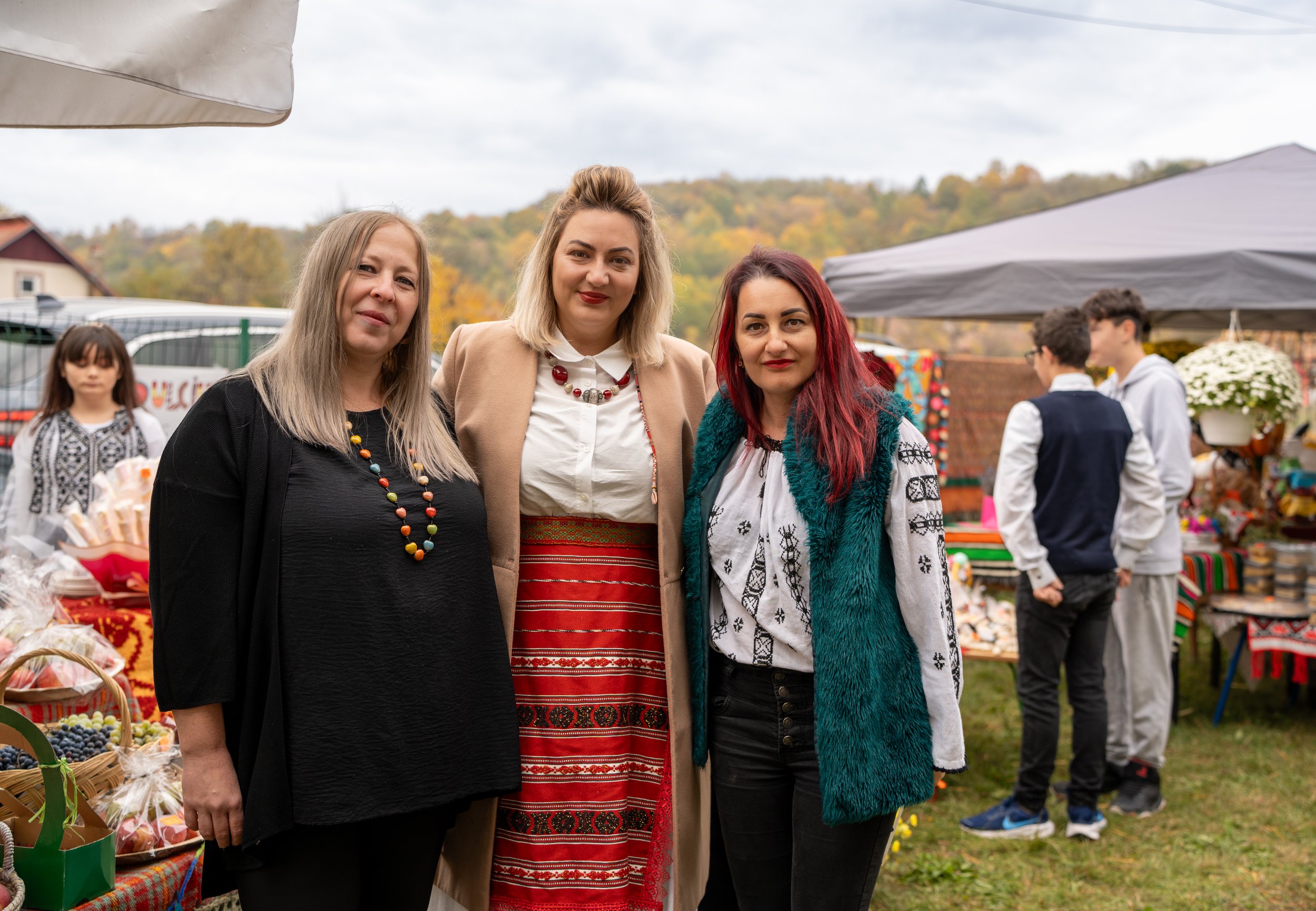 Three women standing together at an outdoor market with stalls and other people in the background, with trees and hills.