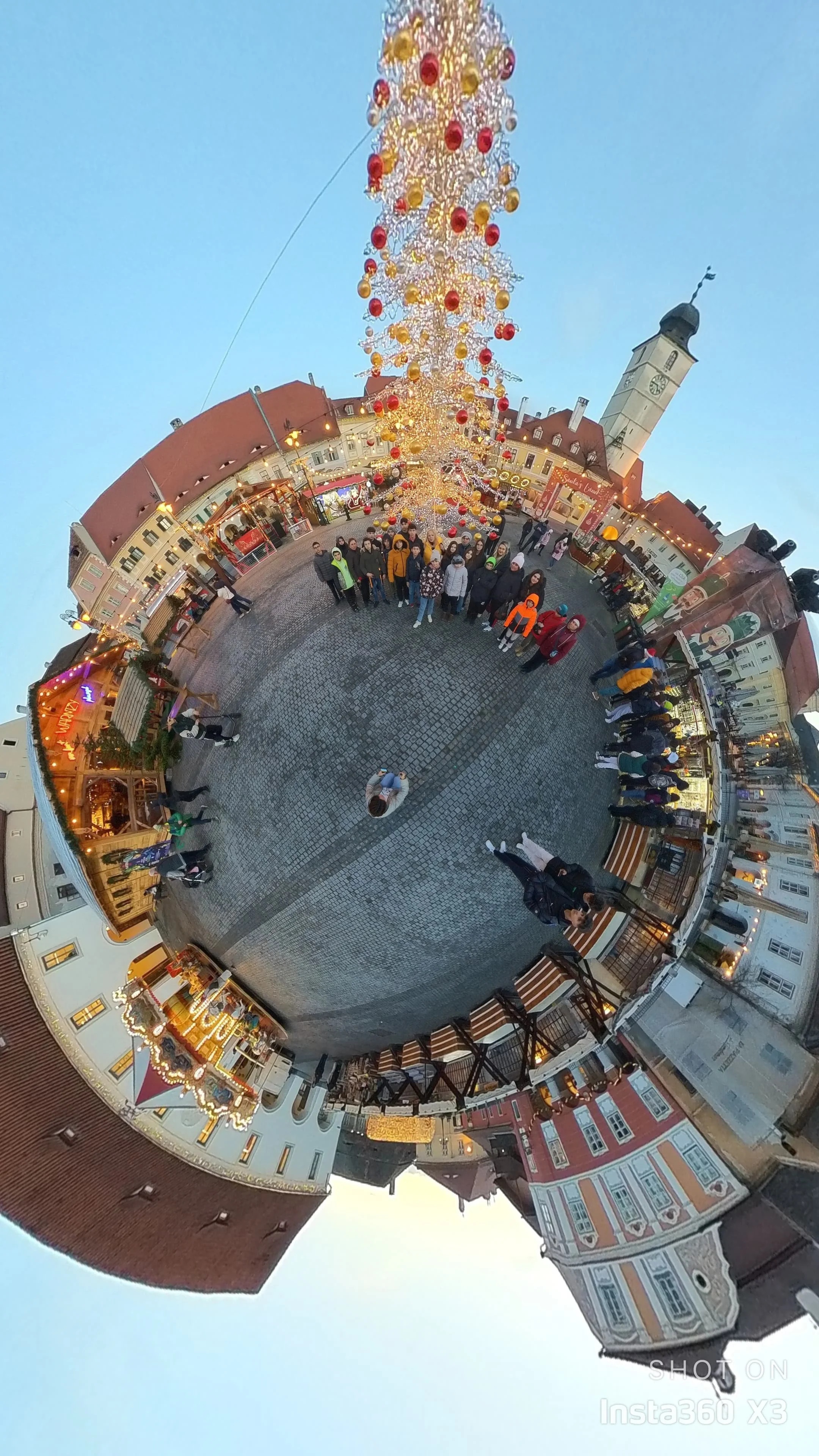 A holiday scene in a town square with a tall Christmas tree decorated with red and yellow ornaments and lights, surrounded by people, buildings, and a clock tower with a spire. The image is captured with a fisheye lens, creating a circular panoramic