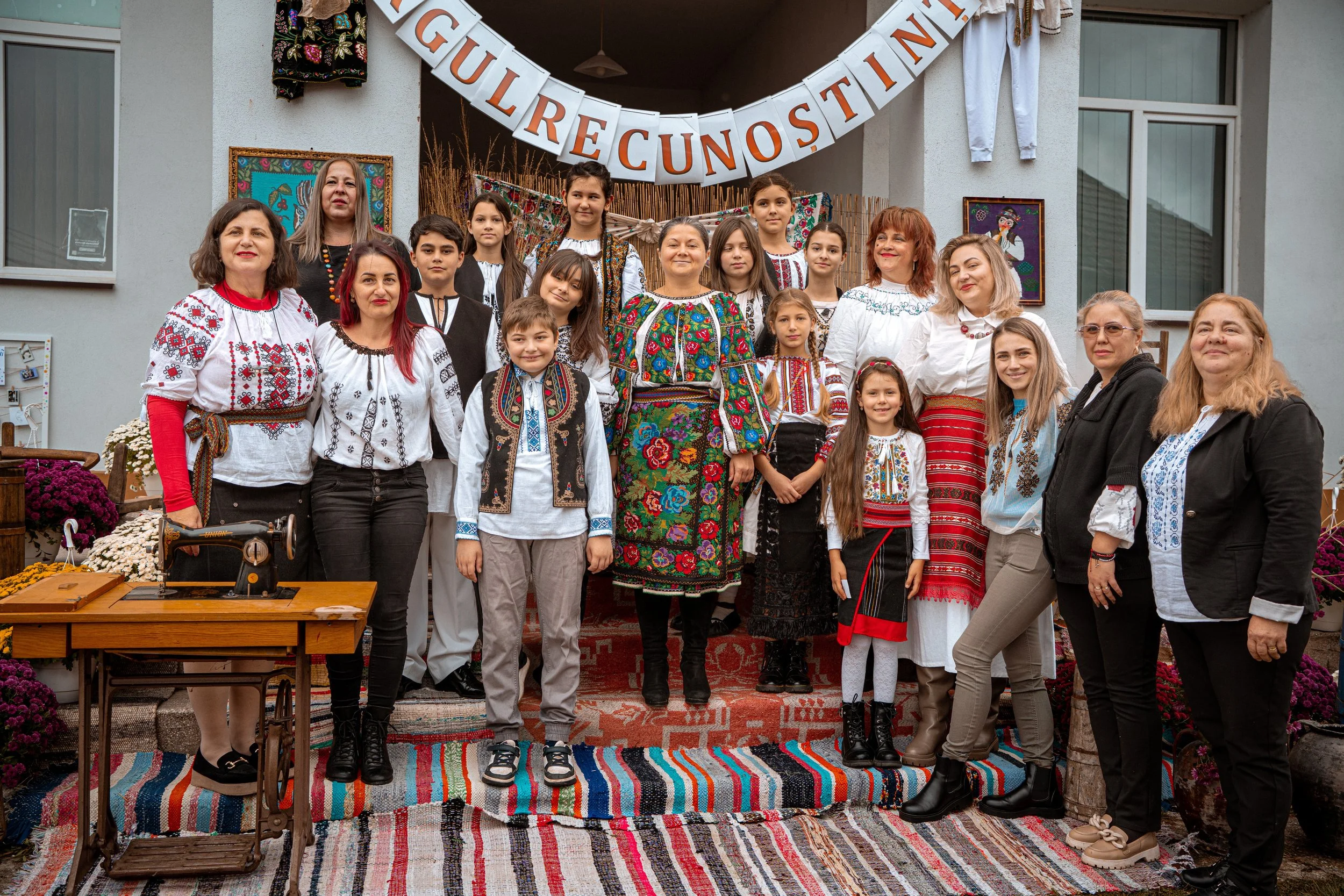 Group of people in traditional embroidered clothing posing on a decorated outdoor stage with a banner that says "Gulre Cunostinta."