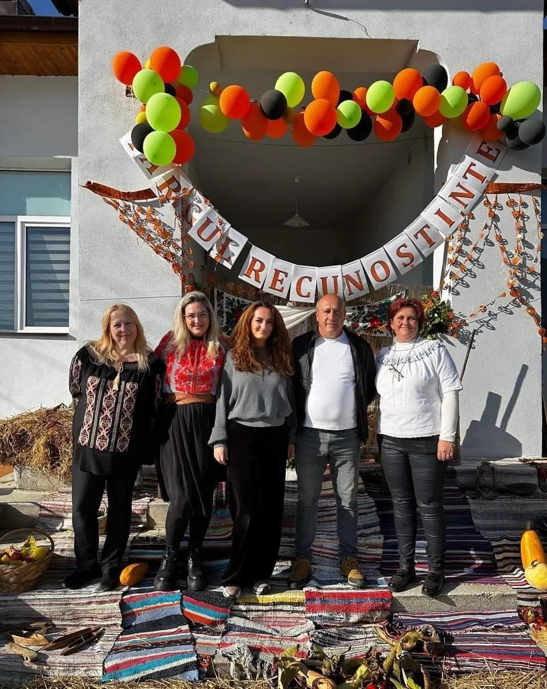 Group of five people standing in front of a decorated outdoor setting with balloons in orange, green, and black, a banner with Romanian text, and traditional woven rugs, suggesting a festive or cultural event.