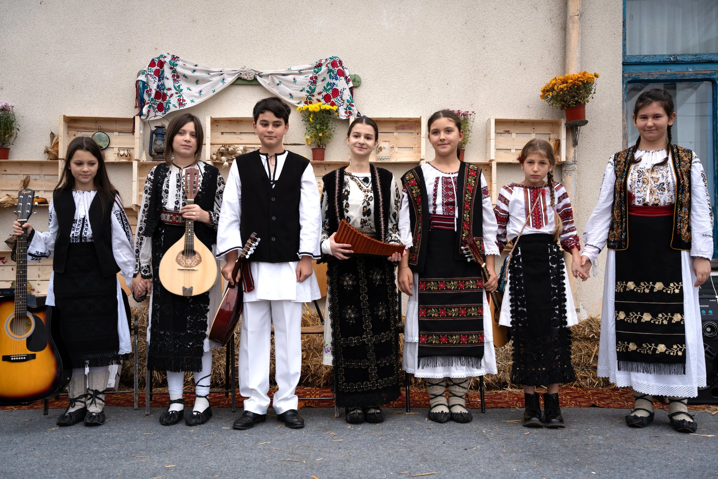 Children dressed in traditional folk costumes standing on stage with musical instruments, holding hands, in front of decorated backdrop with flowers and straw.