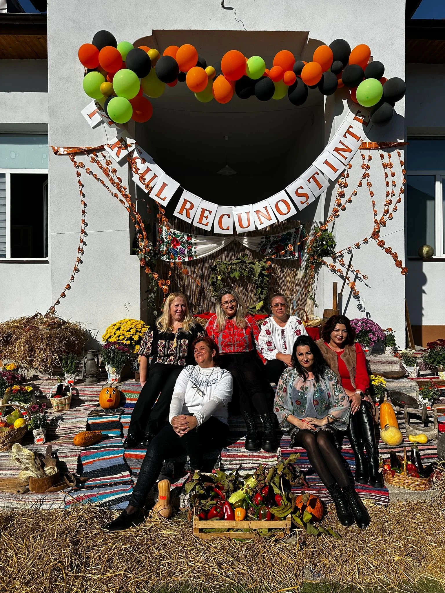Group of six women sitting and posing on a decorated outdoor porch with harvest-themed decorations, pumpkins, and fruits, with a banner that reads "Treiul Recunoștinței" and colorful balloon arrangements above.