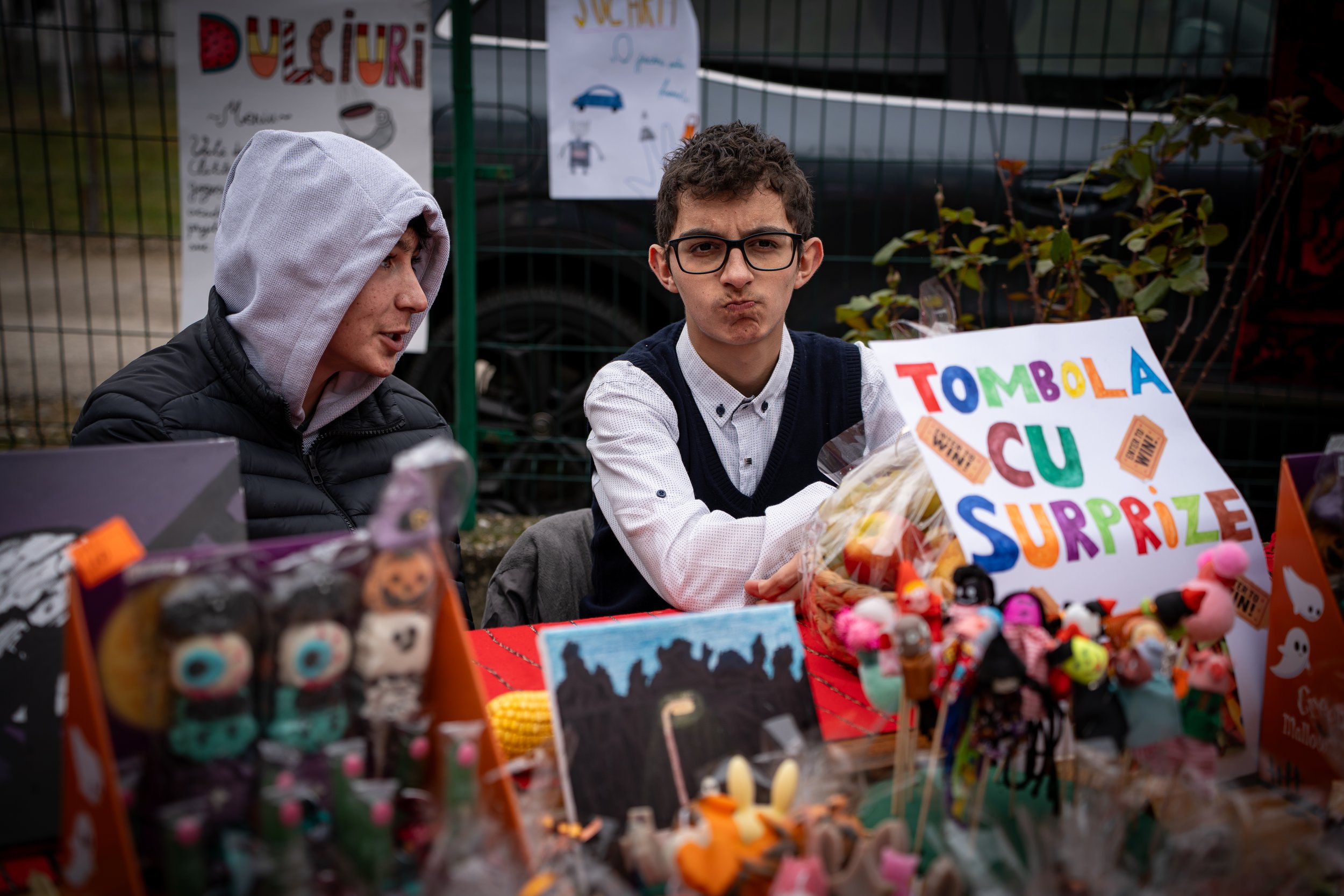 A young man with glasses and a white shirt sitting at a table filled with small plush toys and decorated with a colorful sign that says "TOMBOLA CU SURPRIZE." Next to him, a person in a black jacket and gray hoodie with the hood up talking. The backg