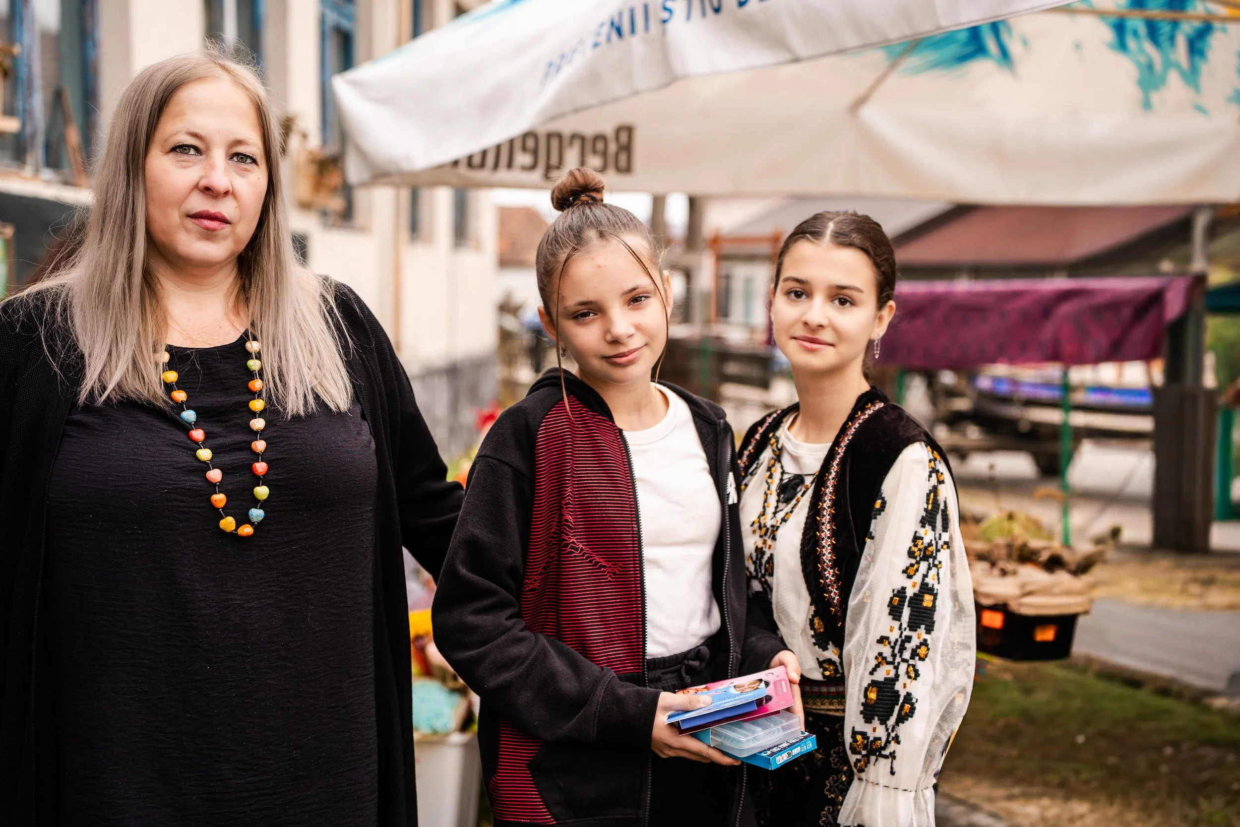 Three females standing outdoors at a market or fair, with tents in the background. The woman on the left has long blonde hair, wears a black dress, and a colorful beaded necklace. The girl in the middle has her hair tied up, wears a black hoodie, and