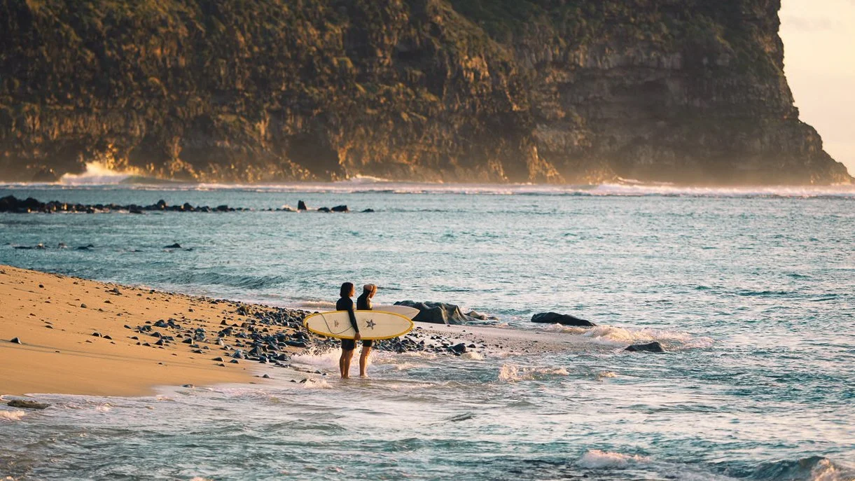 Two surfers on a beach at the base of a ciff, looking out to see