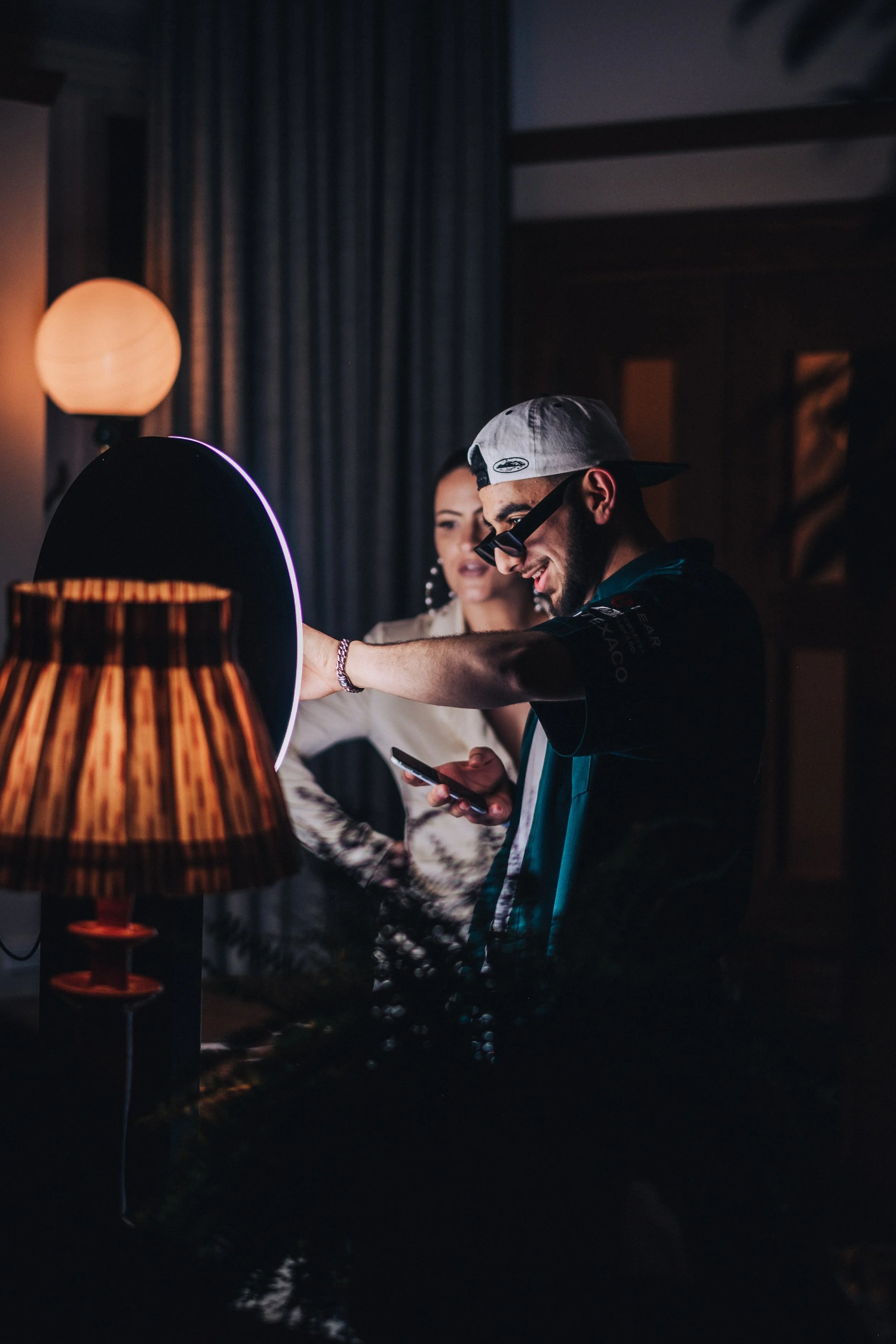 A man wearing sunglasses, a backward cap, and a jacket, smiling while looking at his phone, standing next to a woman with long hair, in a dimly lit room with lamps and dark curtains.