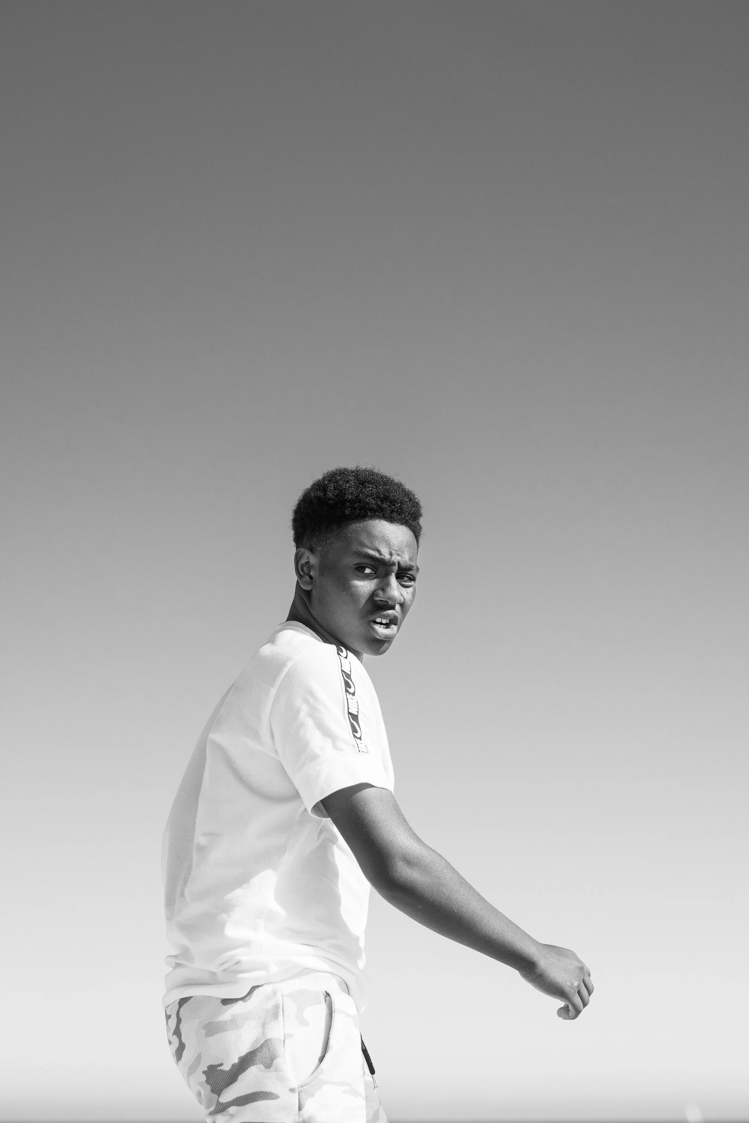 Black and white photo of a young boy with a curious or confused expression, wearing a white t-shirt and camouflage shorts, standing outdoors under a clear sky.