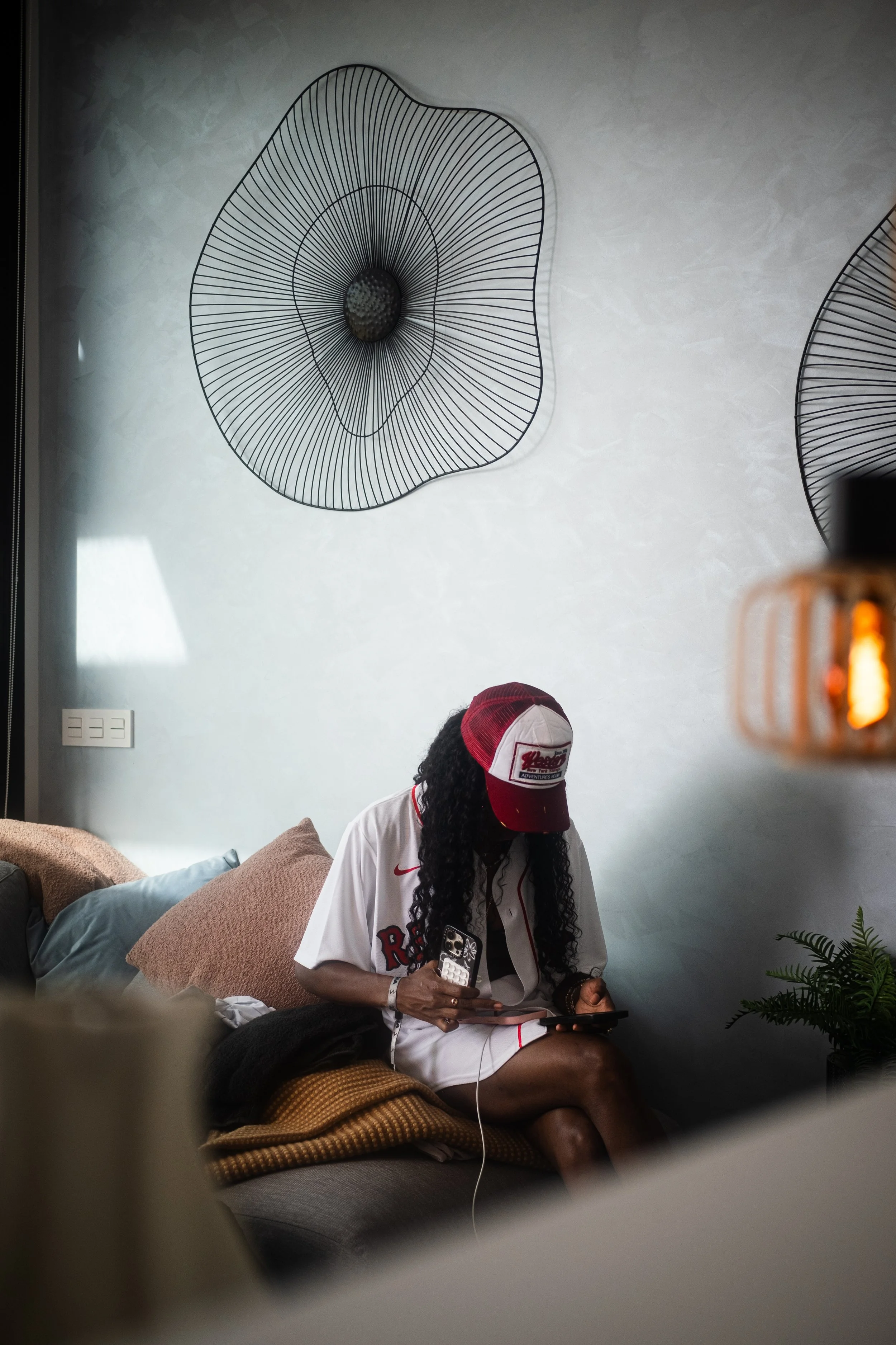 A woman with long curly hair, wearing a red and white baseball cap and a white sports jersey, sitting on a sofa and looking at her phone in a modern living room with wall art and plants.