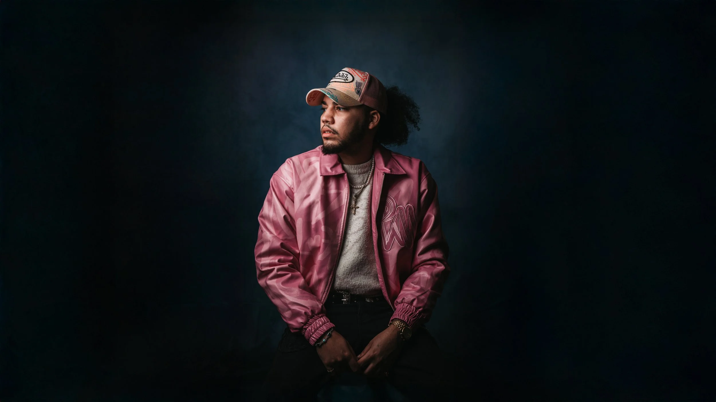 Young man wearing a pink satin jacket, a baseball cap, and jewelry, seated against a dark background.