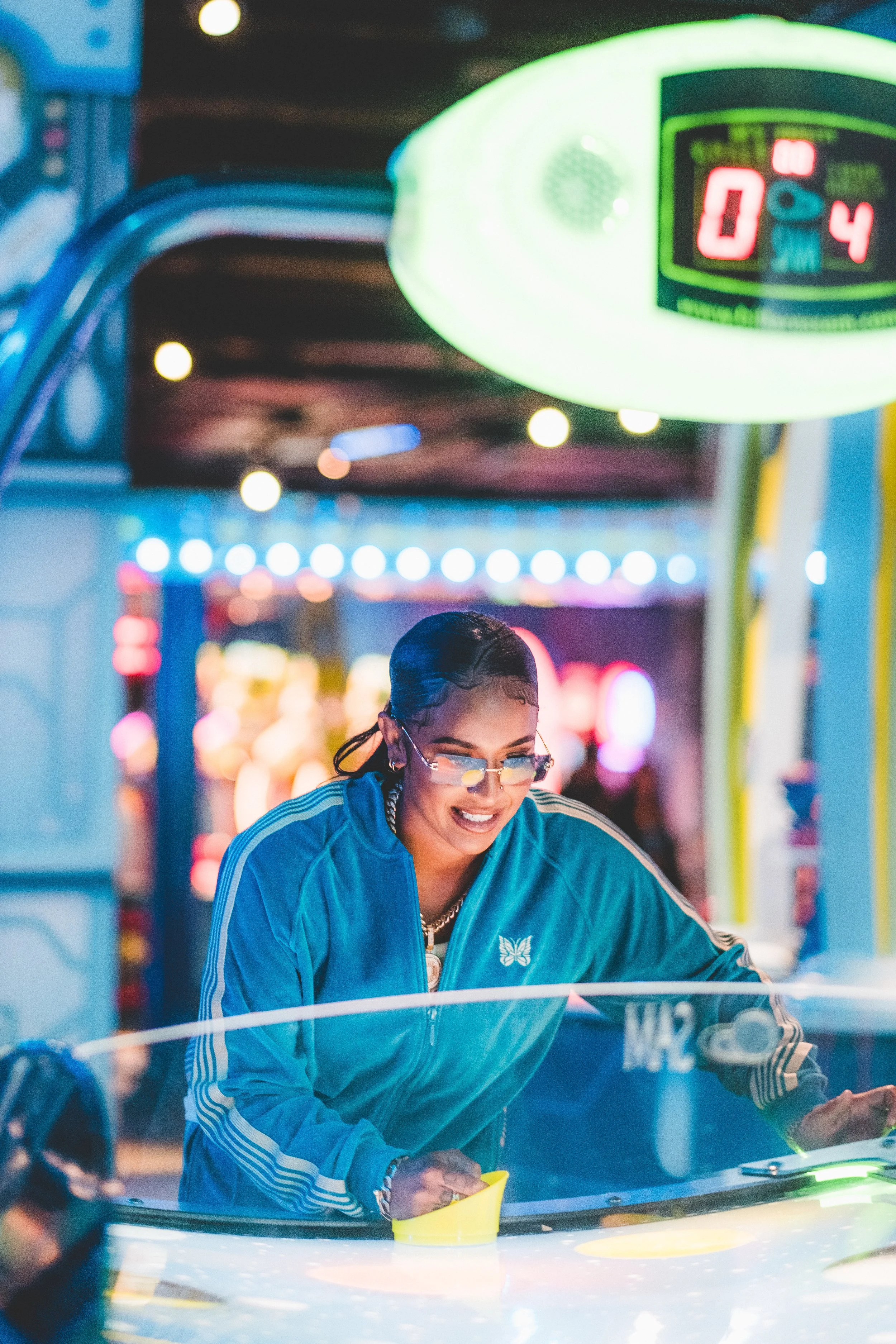 A woman playing air hockey at an arcade, wearing glasses and a blue jacket, with colorful blurred arcade lights in the background.