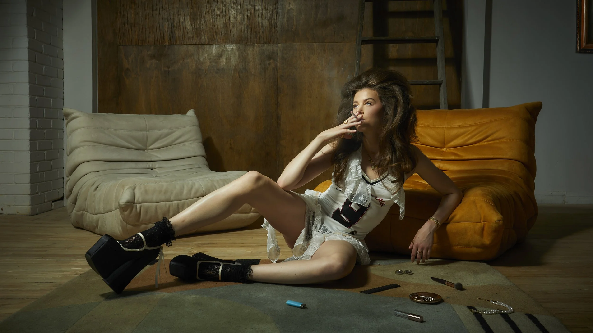 A woman with long, curly brown hair lounging on the floor of a room with wooden and brick walls. She is wearing black platform shoes, lace socks, a white lace dress, and jewelry. Categories of makeup and jewelry accessories are scattered on the floor