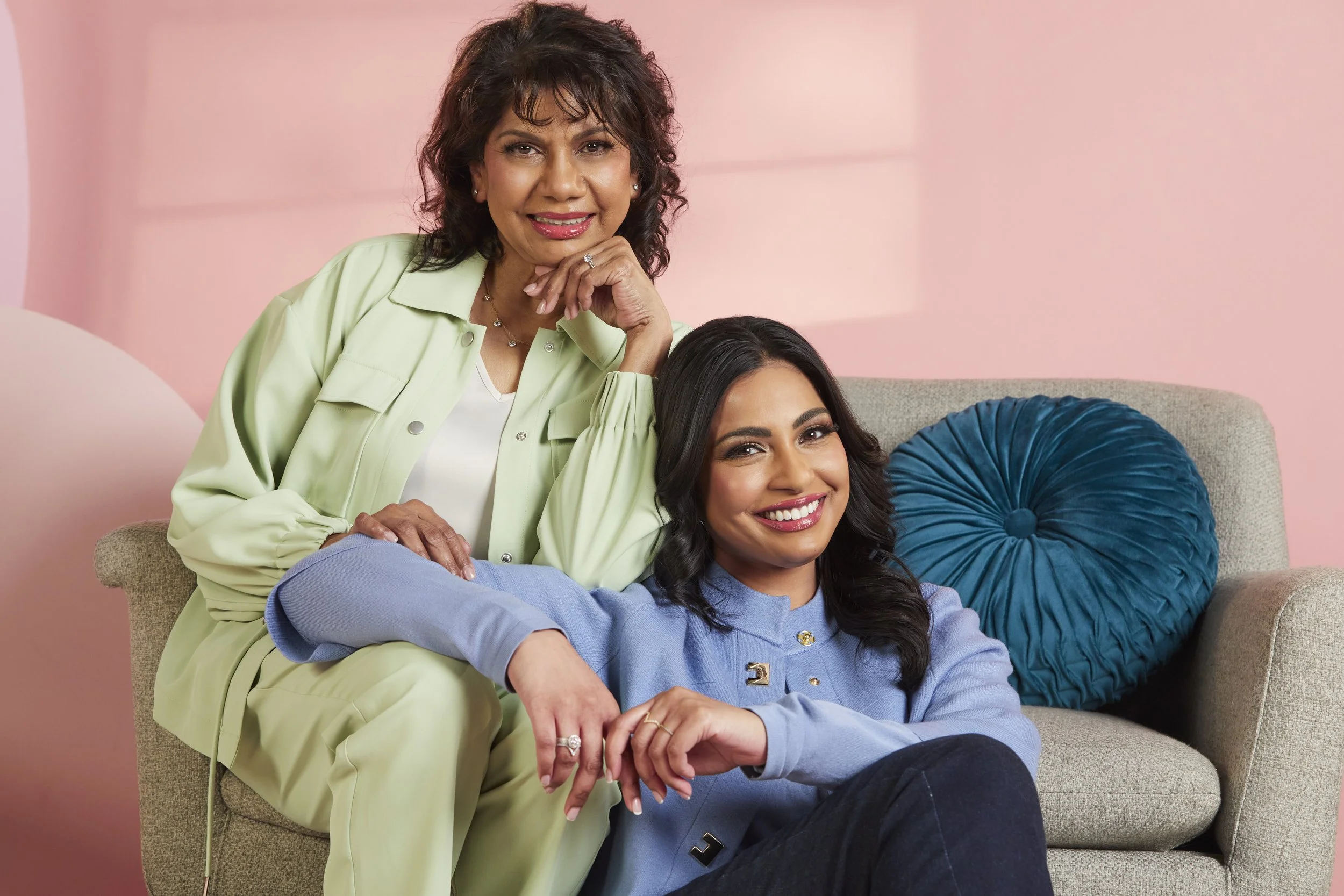Two women, one older and one younger, sitting on a beige sofa with a blue pillow in a room with pink walls. The older woman is standing behind the younger one, resting her hand on the younger woman's shoulder, both smiling at the camera.