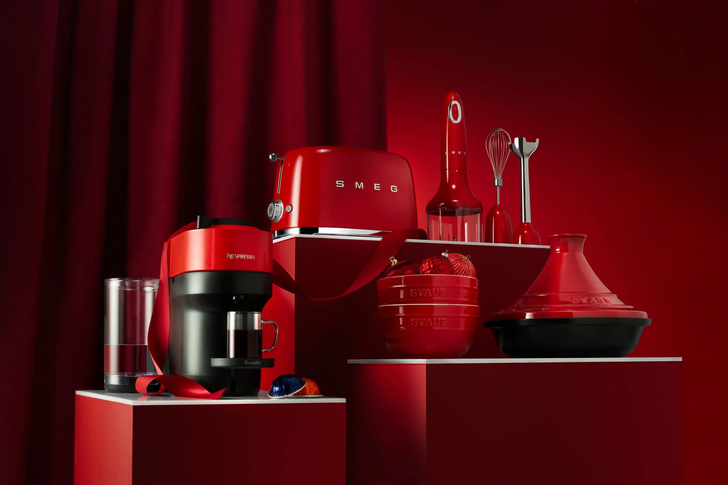 A collection of red and black kitchen appliances and utensils, including a coffee machine, toaster, hand blender, whisk, spatula, and a red bowl, against a red wall and curtain background.