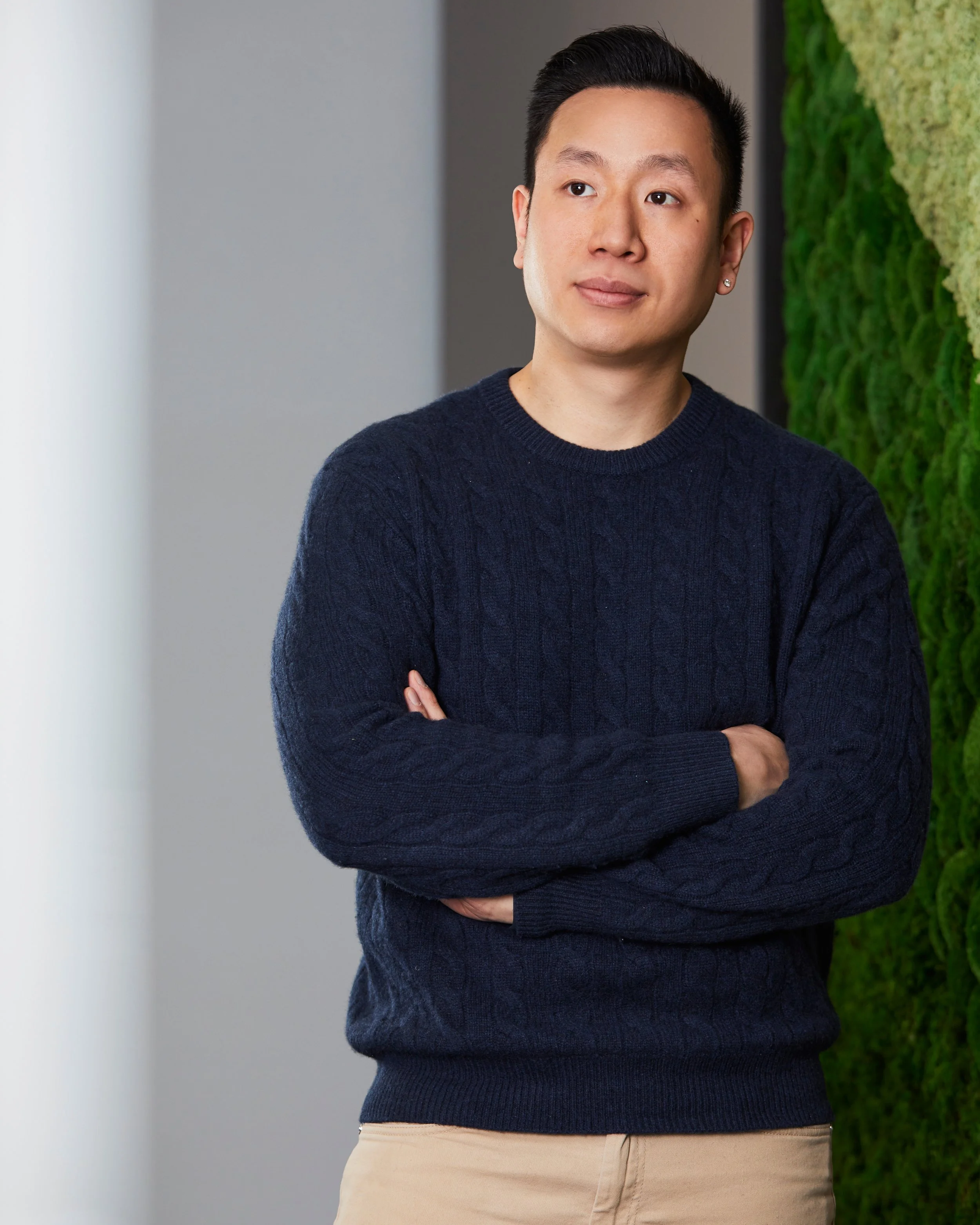 A young man with short black hair, wearing a navy blue sweater and beige pants, standing with arms crossed in front of a green moss wall.