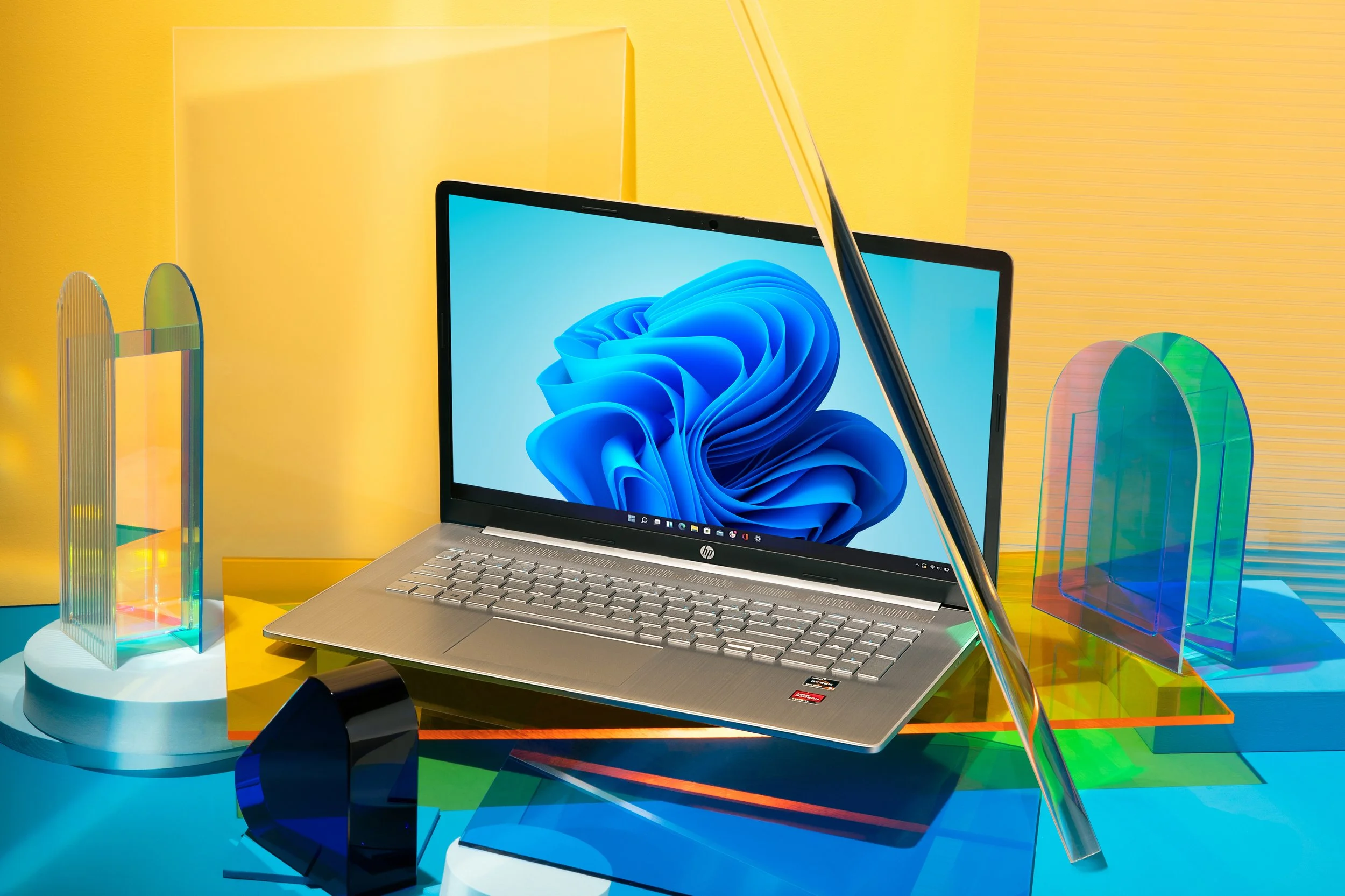 A silver laptop with a blue screen displaying a digital abstract design, surrounded by colorful, translucent, curved acrylic displays on a multicolored table with a yellow background.