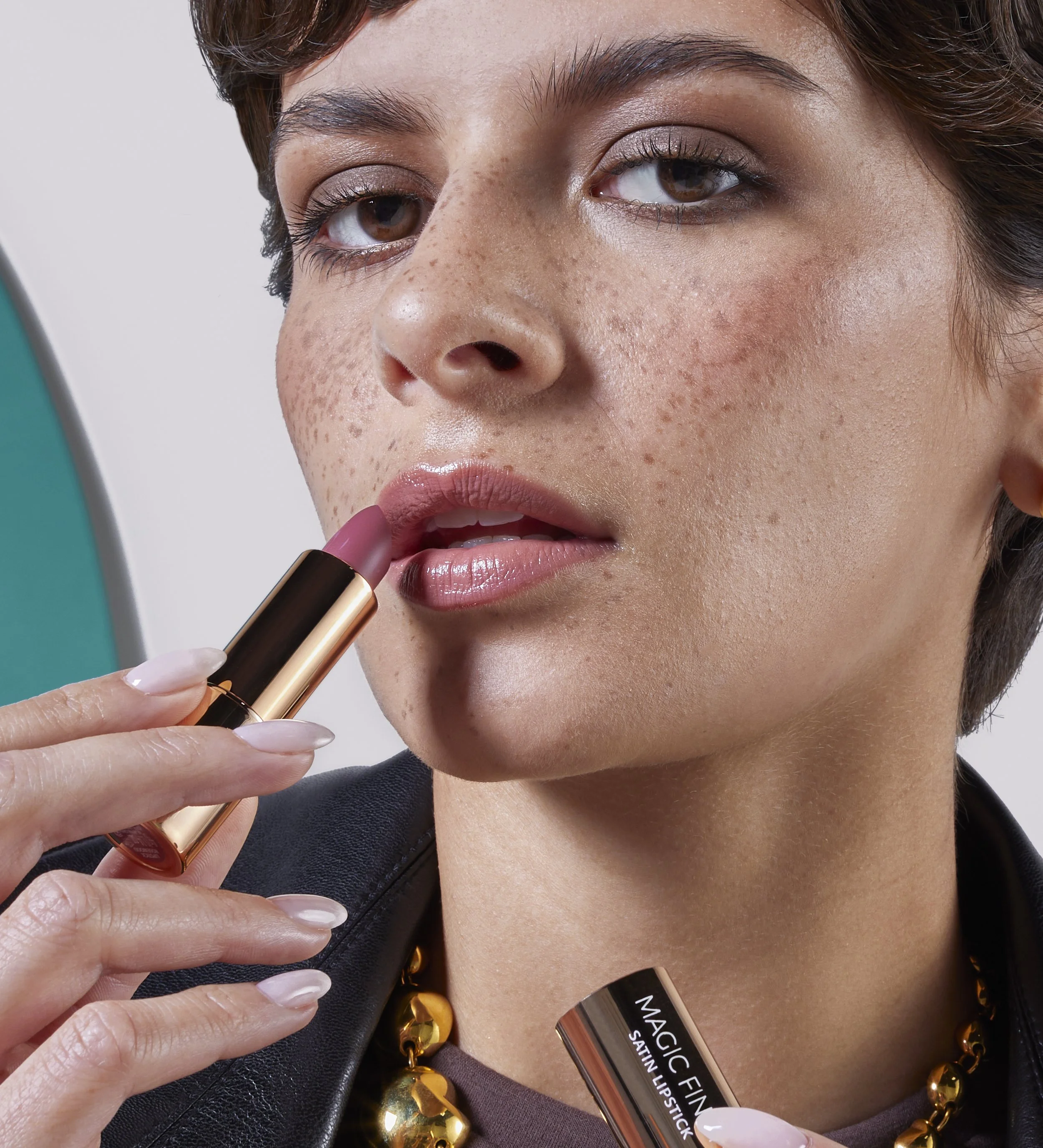 A woman with brown hair, freckles, and brown eyes applying lipstick with gold packaging. She wears natural makeup, a black leather jacket, and a statement gold necklace. She holds a lipstick tube labeled "Magic Finish Satin Lipstick" near her lips, w