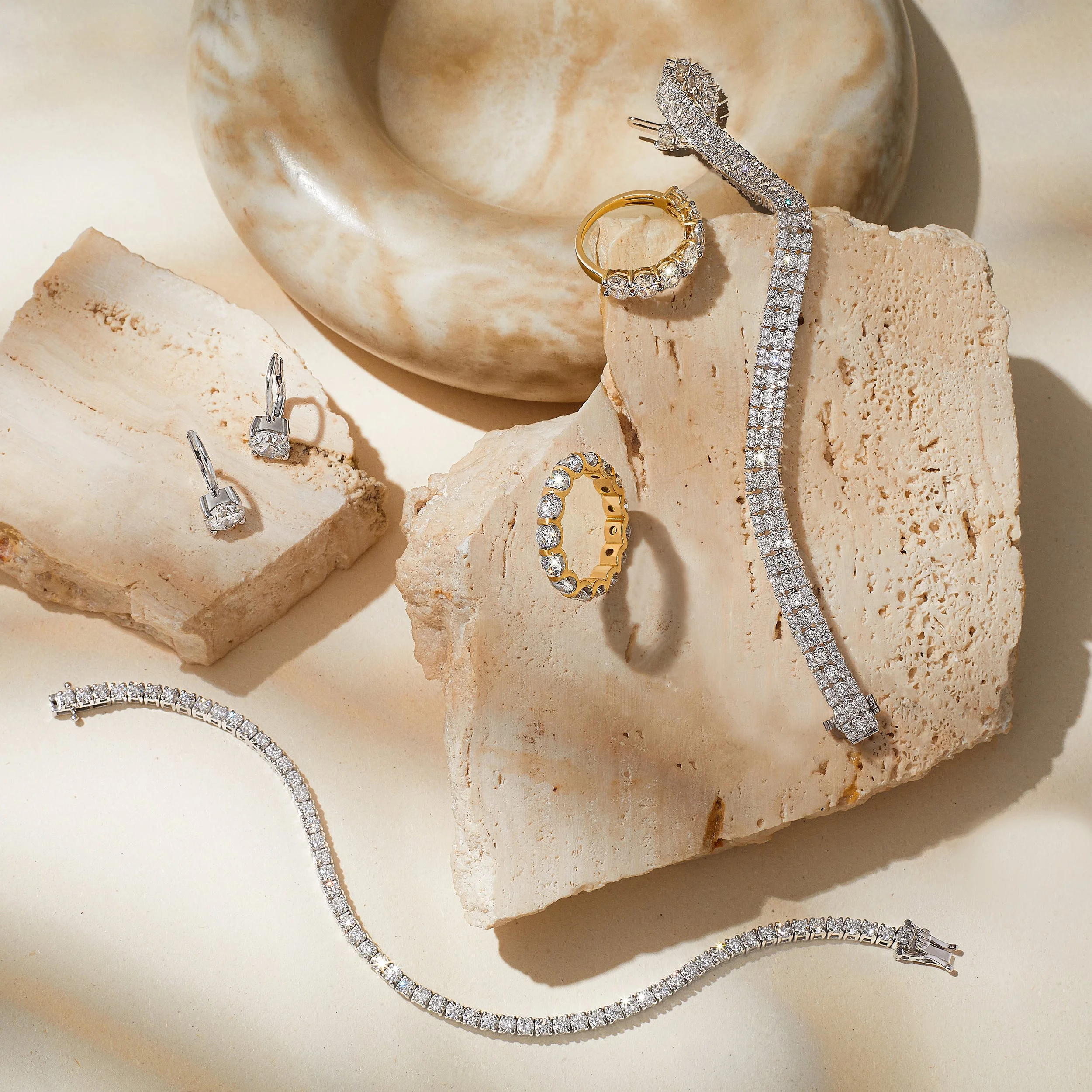 Various jewelry pieces including diamond earrings, rings, and bracelets displayed on beige textured stones and a cream-colored surface.