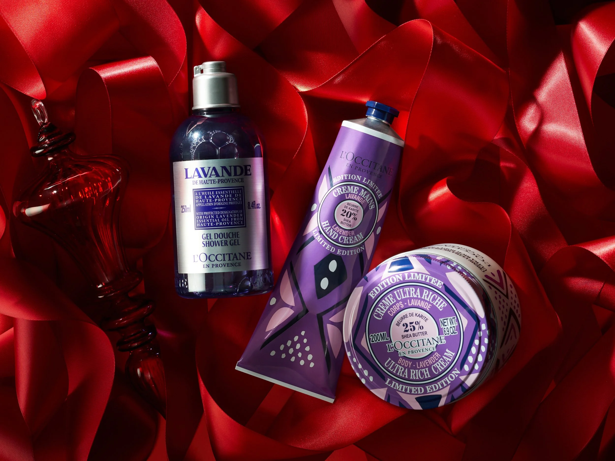 A collection of lavender-scented skincare products on red satin fabric, including shower gel, hand cream, body cream, and a small glass perfume bottle.