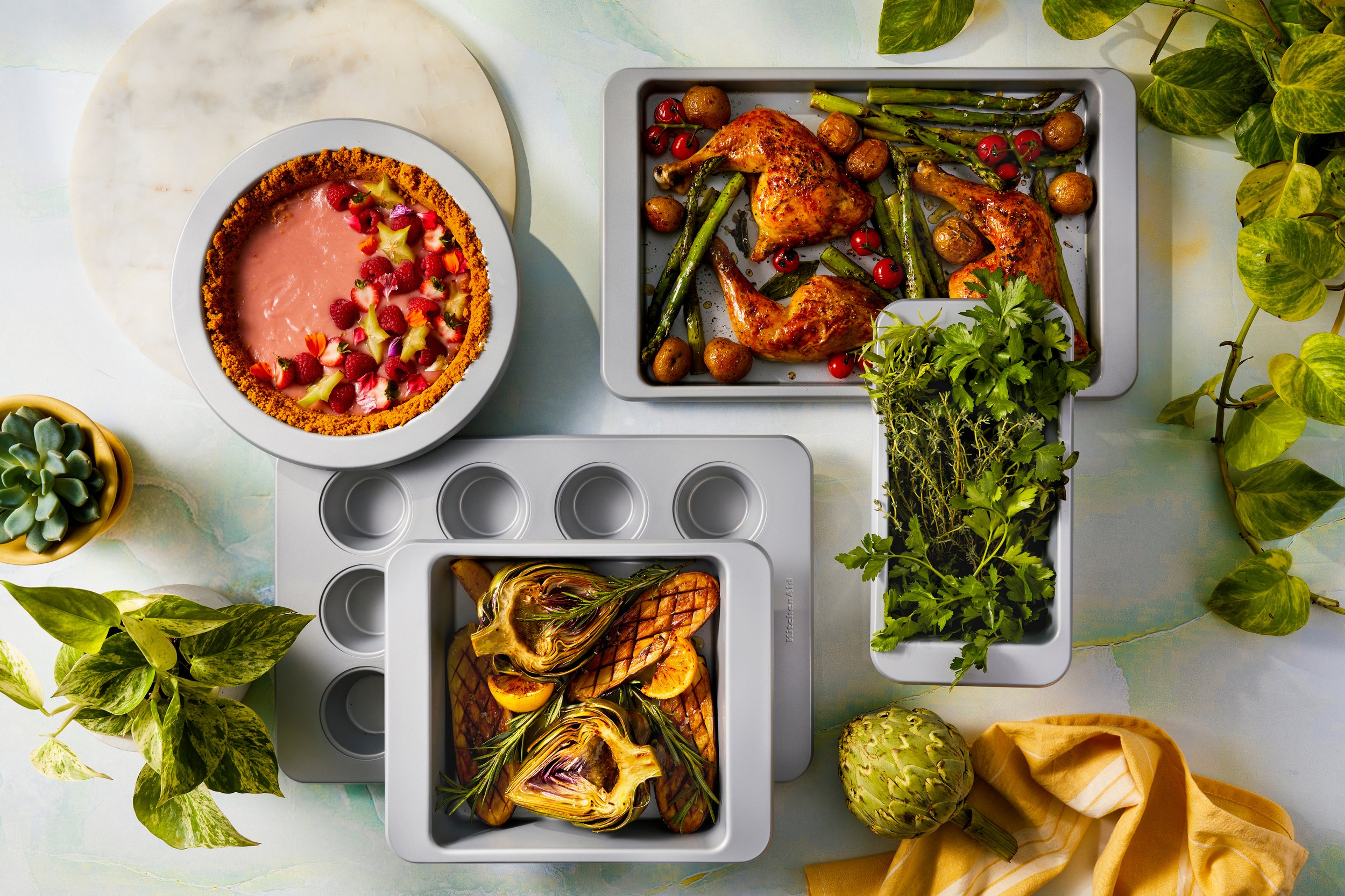 A flat lay of a meal including roasted chicken with vegetables, grilled eggplants with herbs, a berry tart, and various potted plants on a light table.