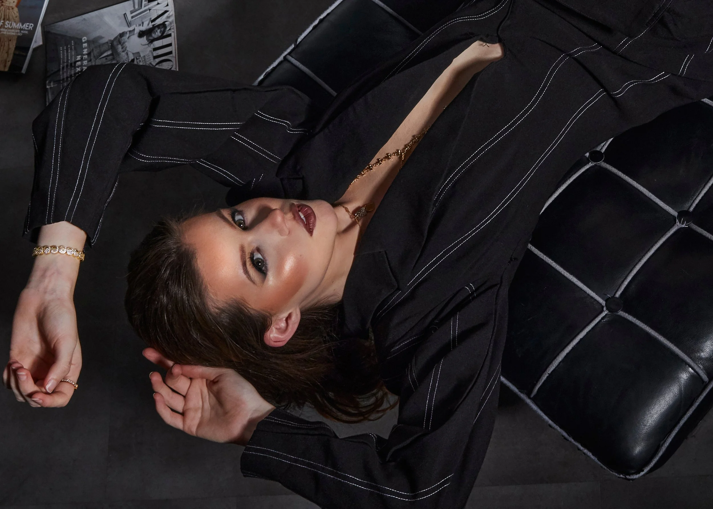 A woman lying on a black leather tufted sofa, wearing a black top with white stitching, gold jewelry, makeup with highlighted cheekbones and dark lipstick, with magazines visible in the background.