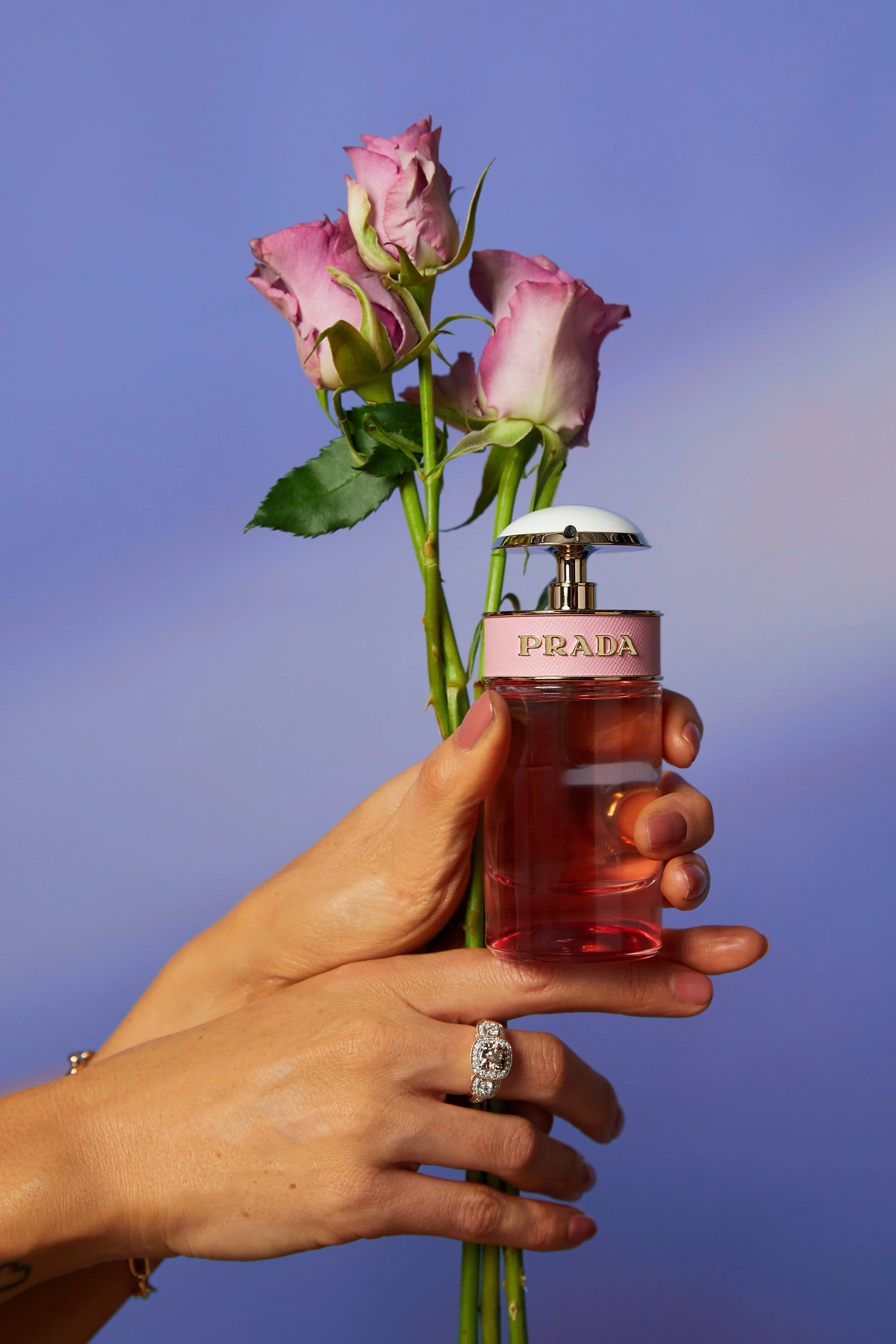 A hand with a ring holds a pink PRADA perfume bottle, which is positioned in front of pink roses against a blue background.