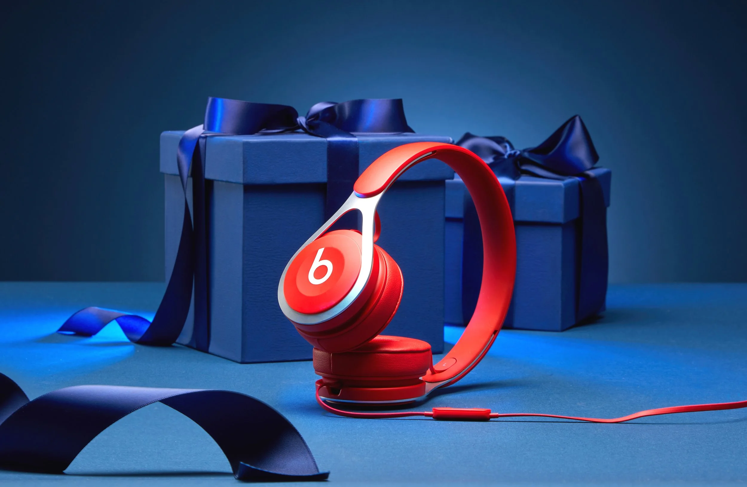Red Beats headphones placed on a blue surface, with two blue wrapped gift boxes with ribbons in the background.