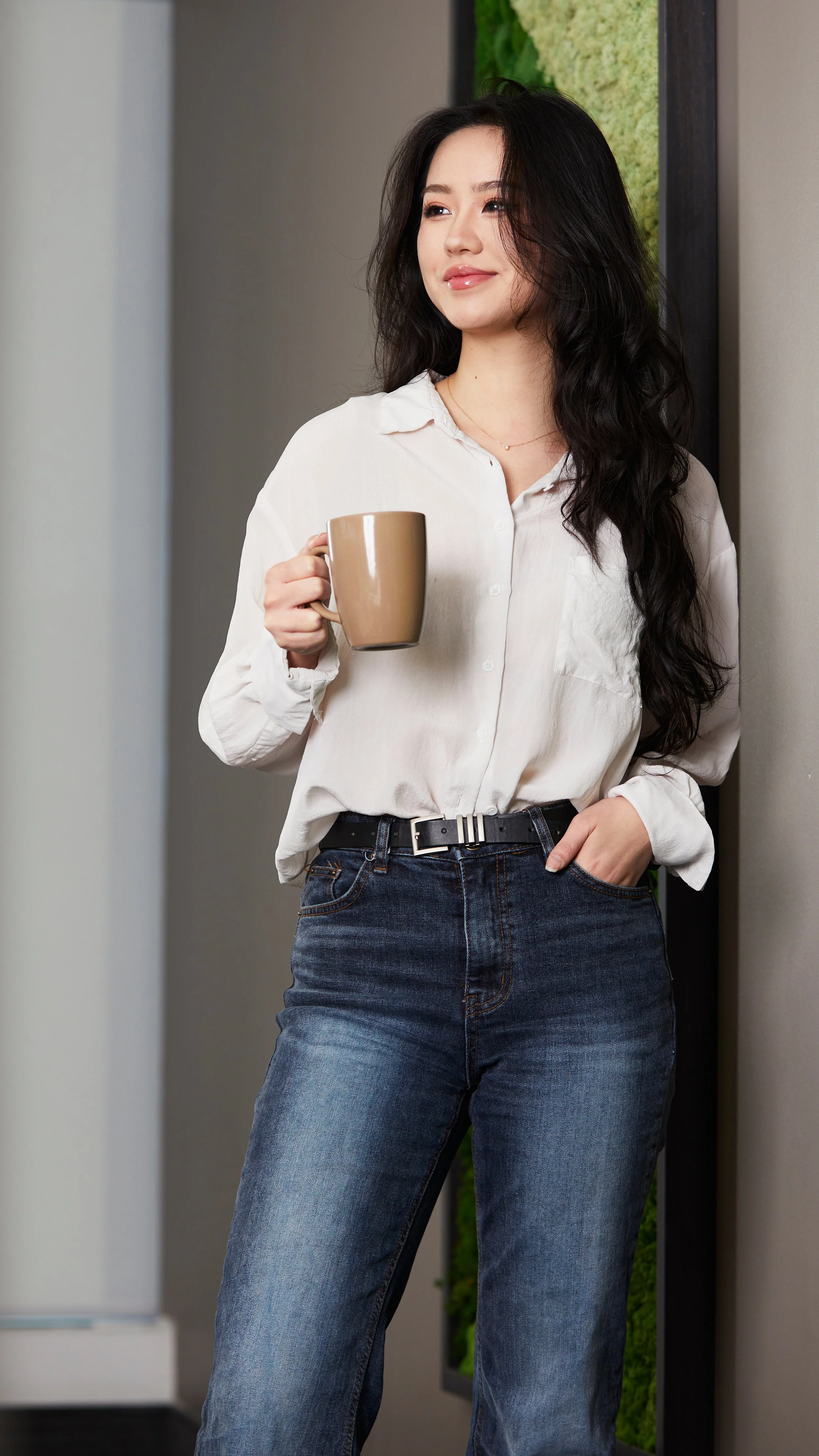 Young woman with long dark hair, wearing a white button-up shirt and blue jeans, holding a beige coffee mug, standing indoors near a wall with a green decorative panel.