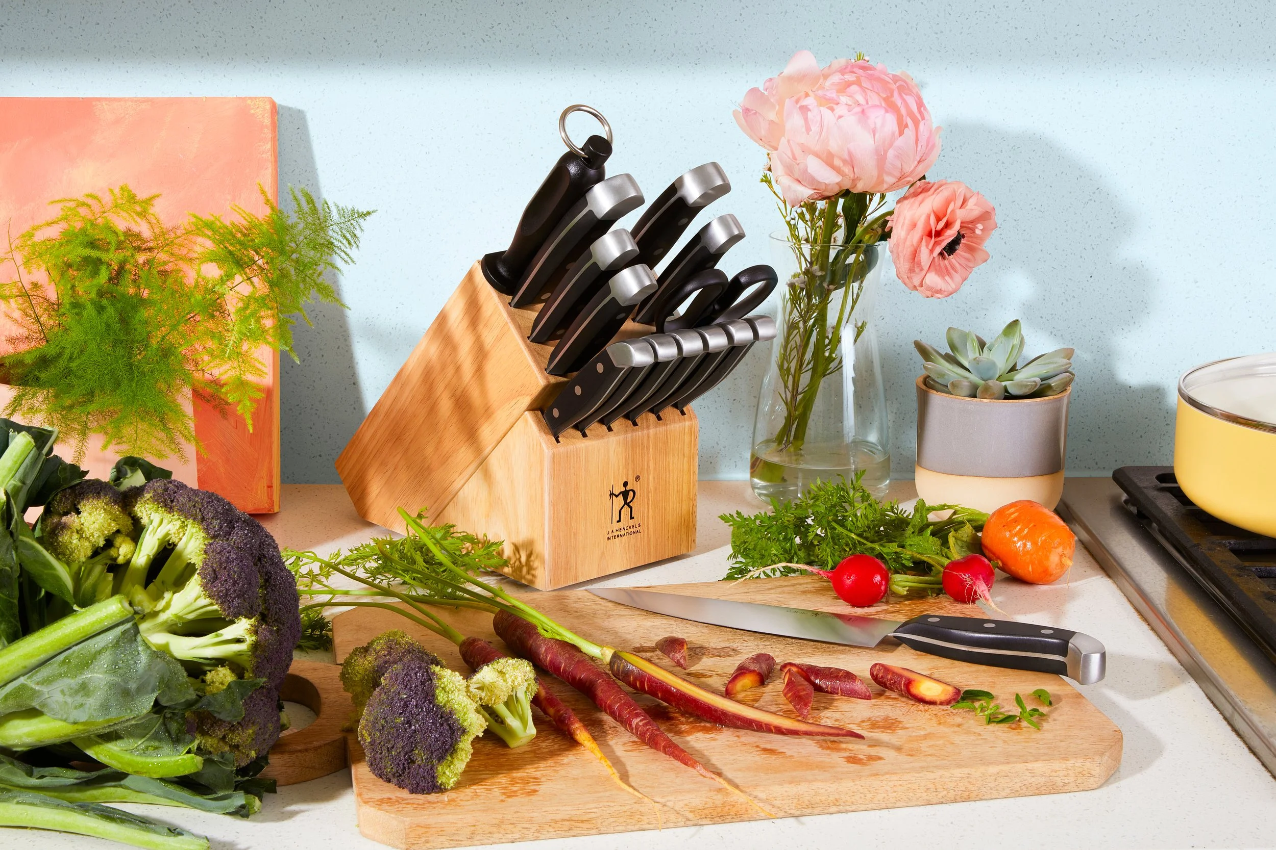 Kitchen countertop with fresh vegetables, a knife, a wooden knife block with knives, a vase with pink flowers, a potted succulent, a pot on the stove, and a cutting board with vegetables.