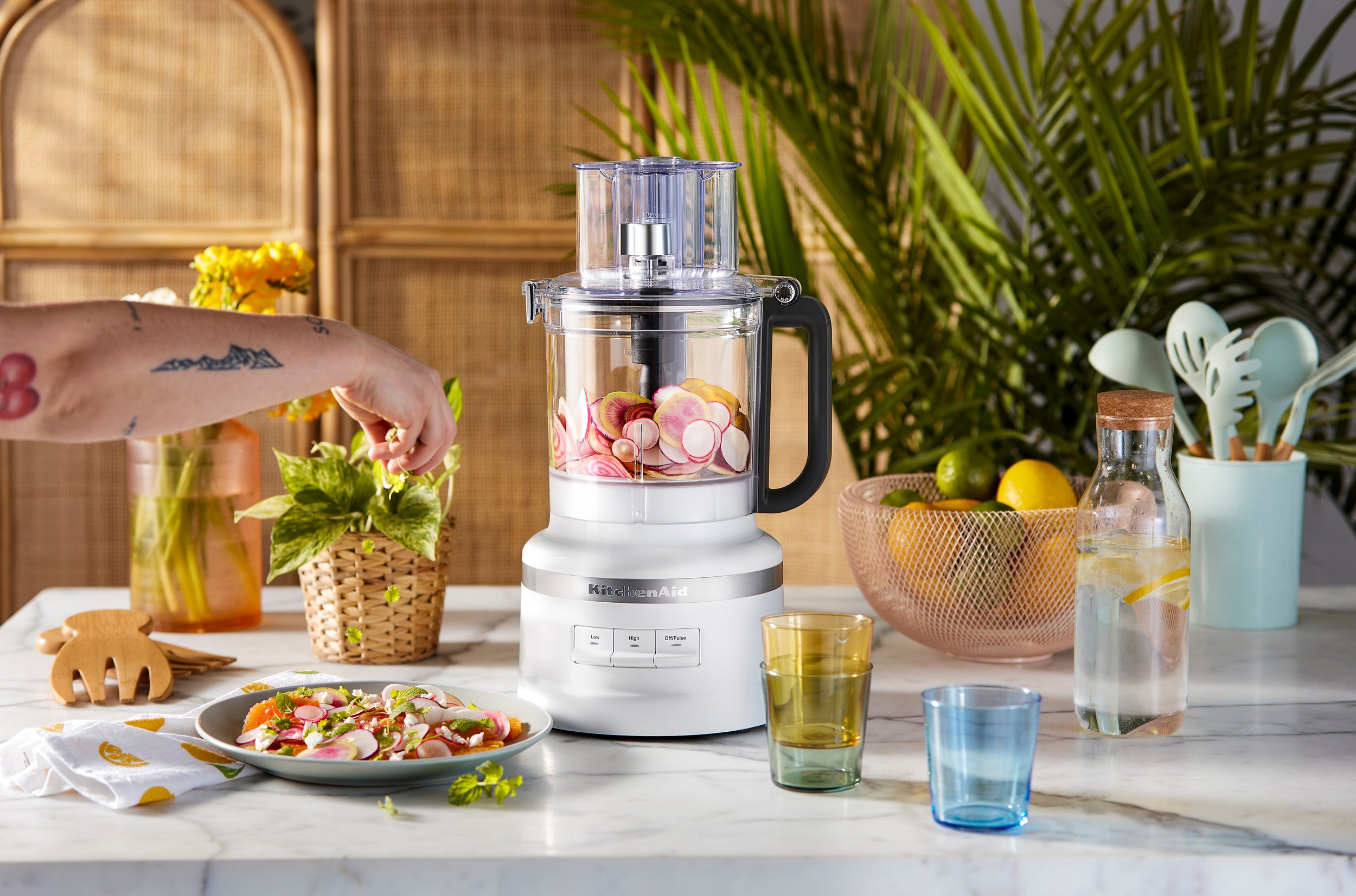 A kitchen scene with a food processor blending radishes, a plate of sliced radishes and herbs, a potted plant, a bowl of limes and lemons, a pitcher of water with lemon slices, colorful glasses, and kitchen utensils on a marble countertop.