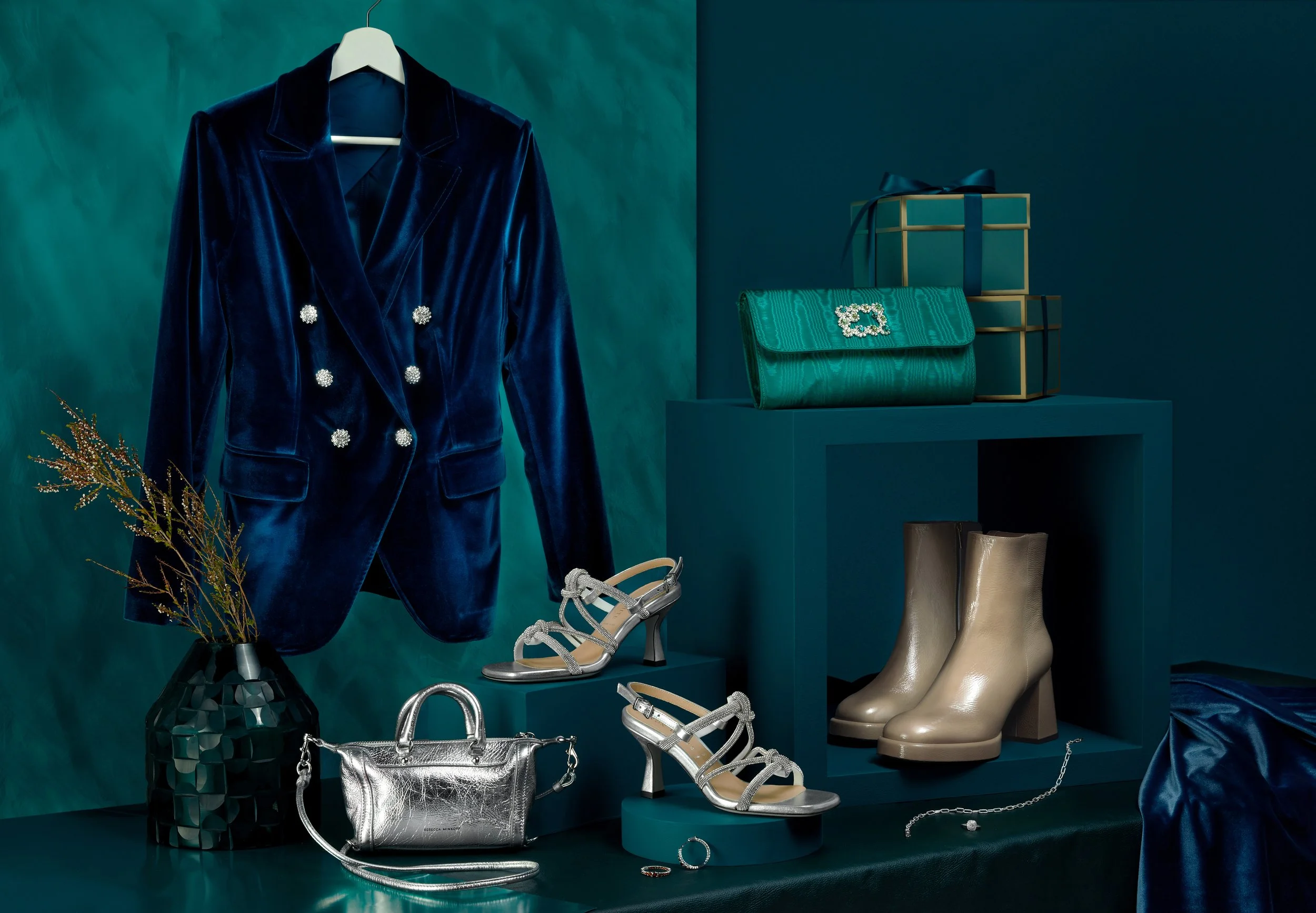 Display of fashion accessories including a blue velvet blazer, silver high-heeled sandals, a metallic silver purse, beige ankle boots, a green textured clutch, layered boxes with a ribbon, jewelry, and a black vase with plants on a teal background.