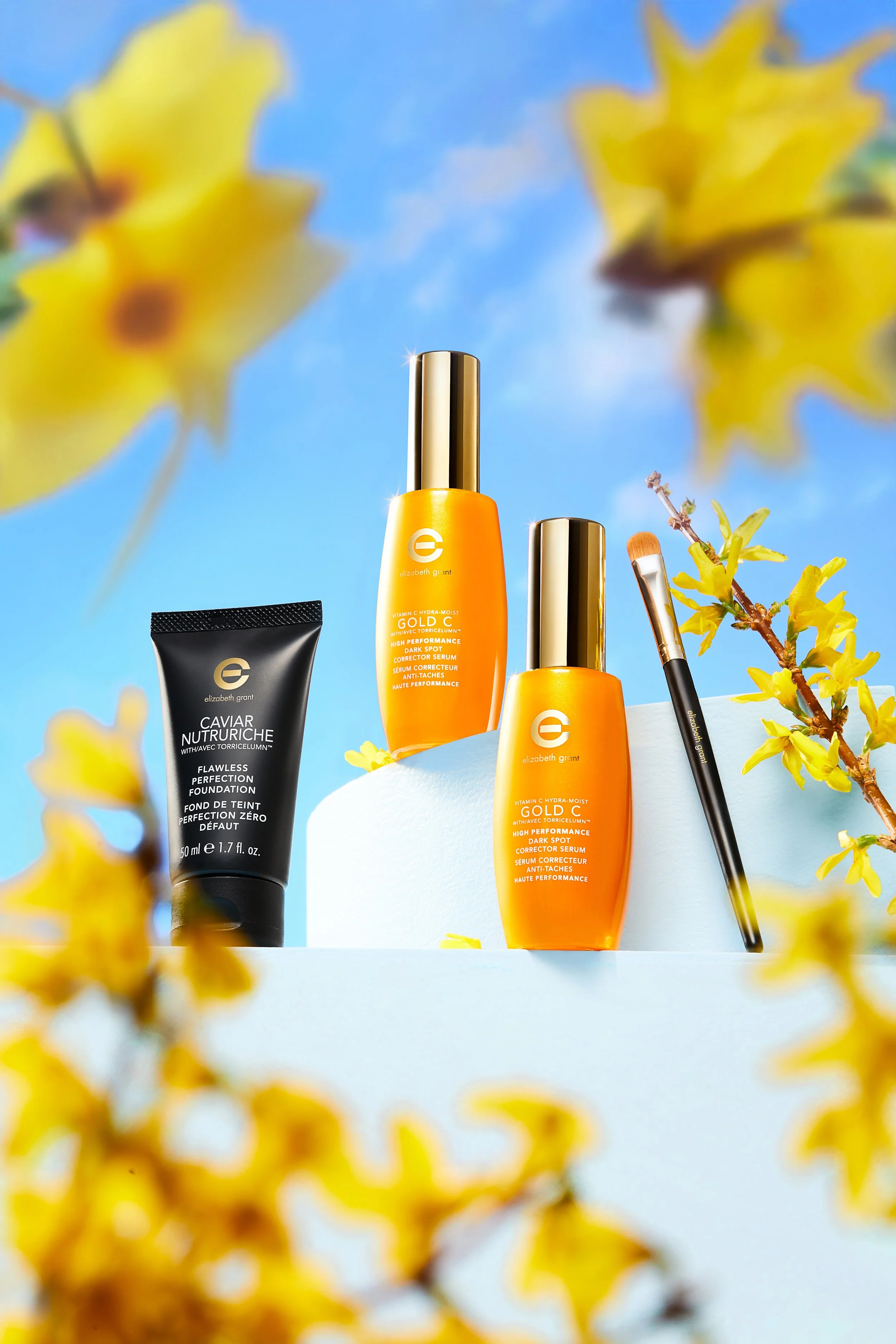 Beauty products, including two Elizabeth Grant bottles, a black tube, and a makeup brush, arranged outdoors on a white surface with yellow flowers and a blue sky in the background.