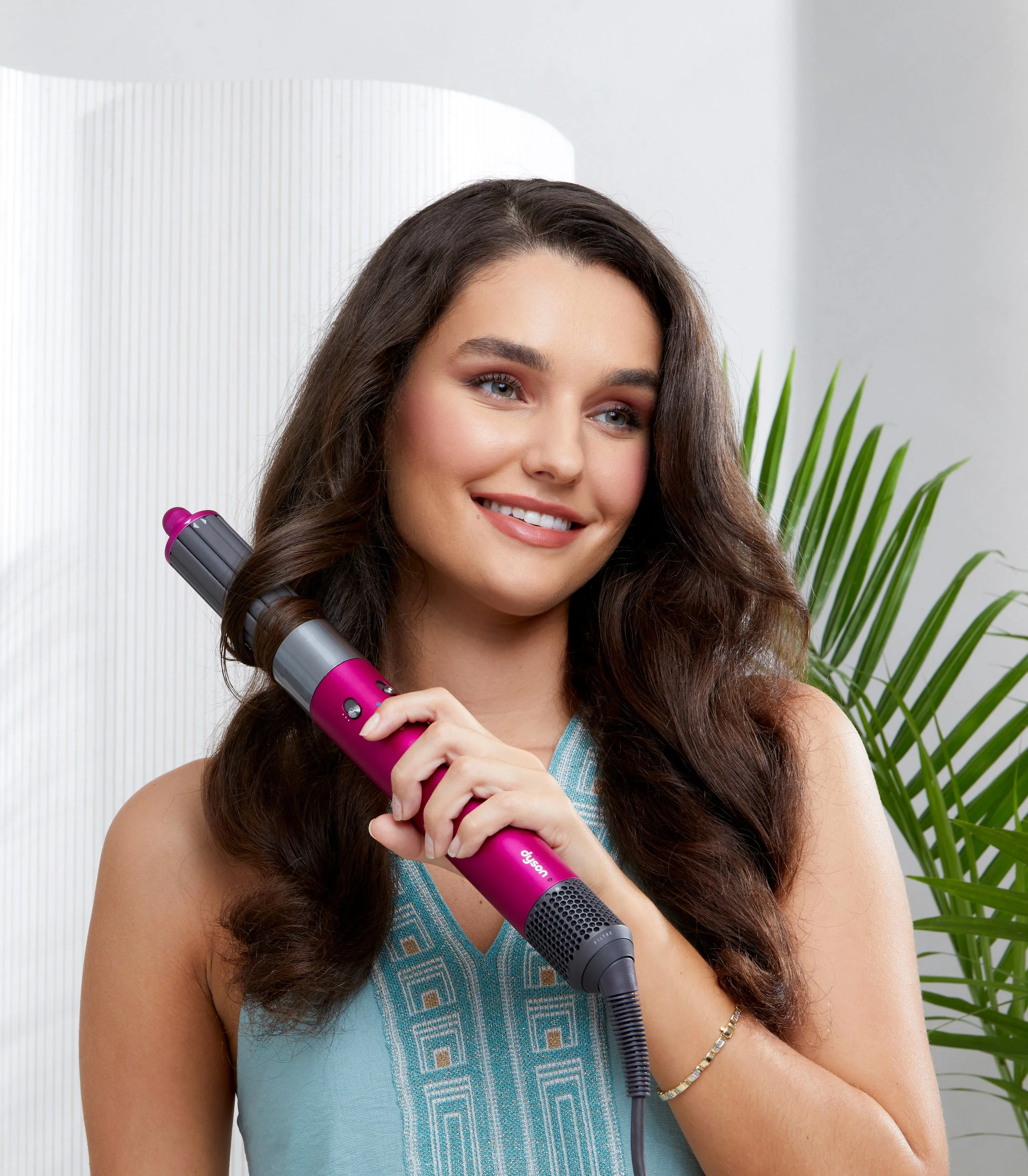 Woman with long wavy brown hair using a pink and black curling iron while smiling.