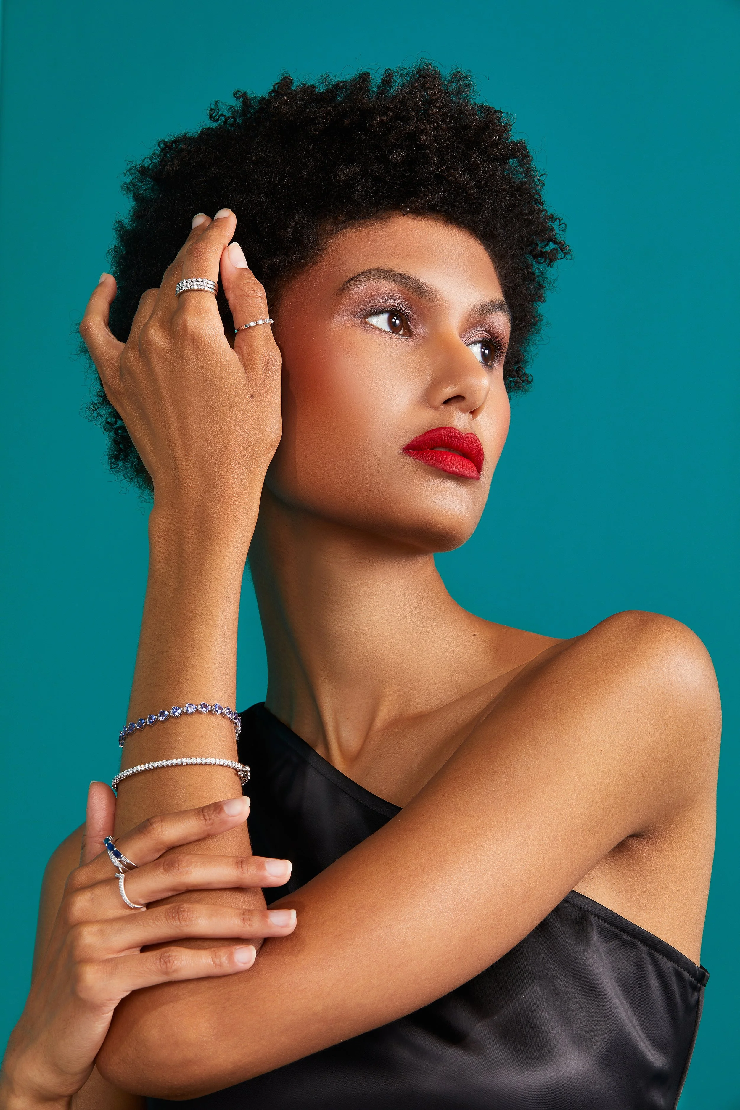 A woman with short curly hair wearing red lipstick and red eyeshadow, posing against a teal background. She is dressed in a black satin top and displaying various rings, bracelets, and a necklace.
