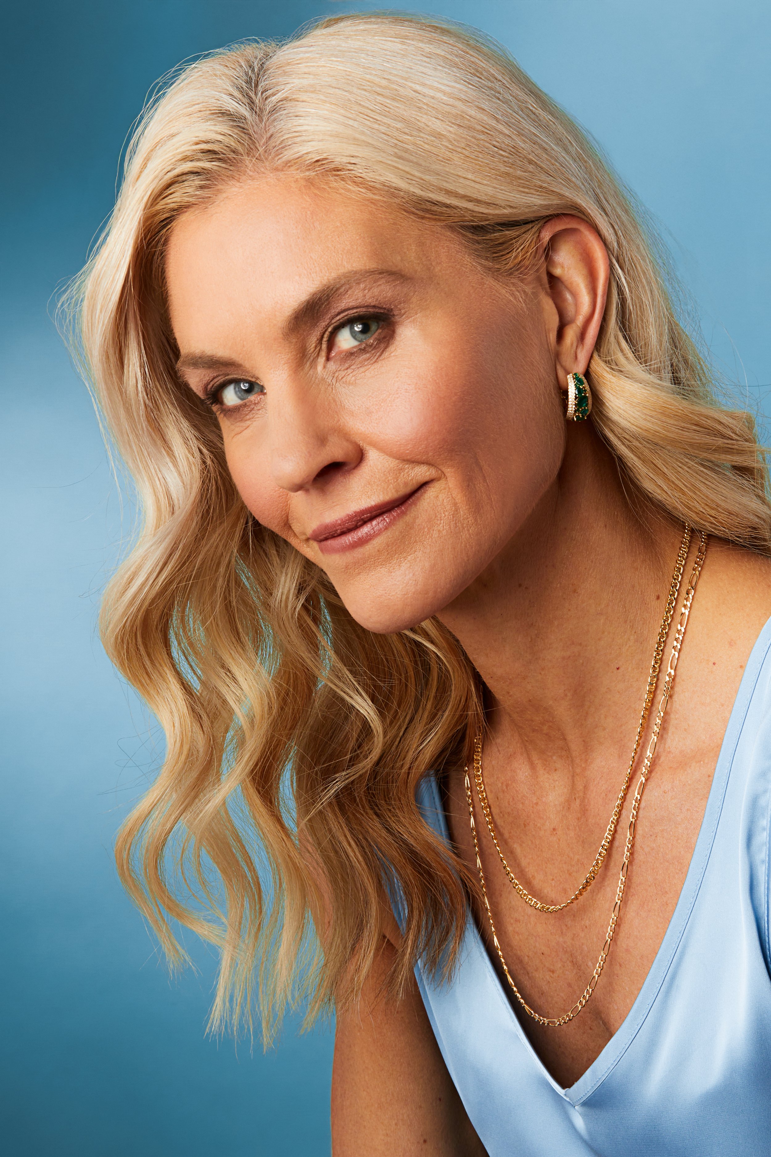 Close-up portrait of a middle-aged woman with blonde hair styled in loose waves, wearing a light blue top, gold jewelry, and small earrings, smiling softly against a blue background.