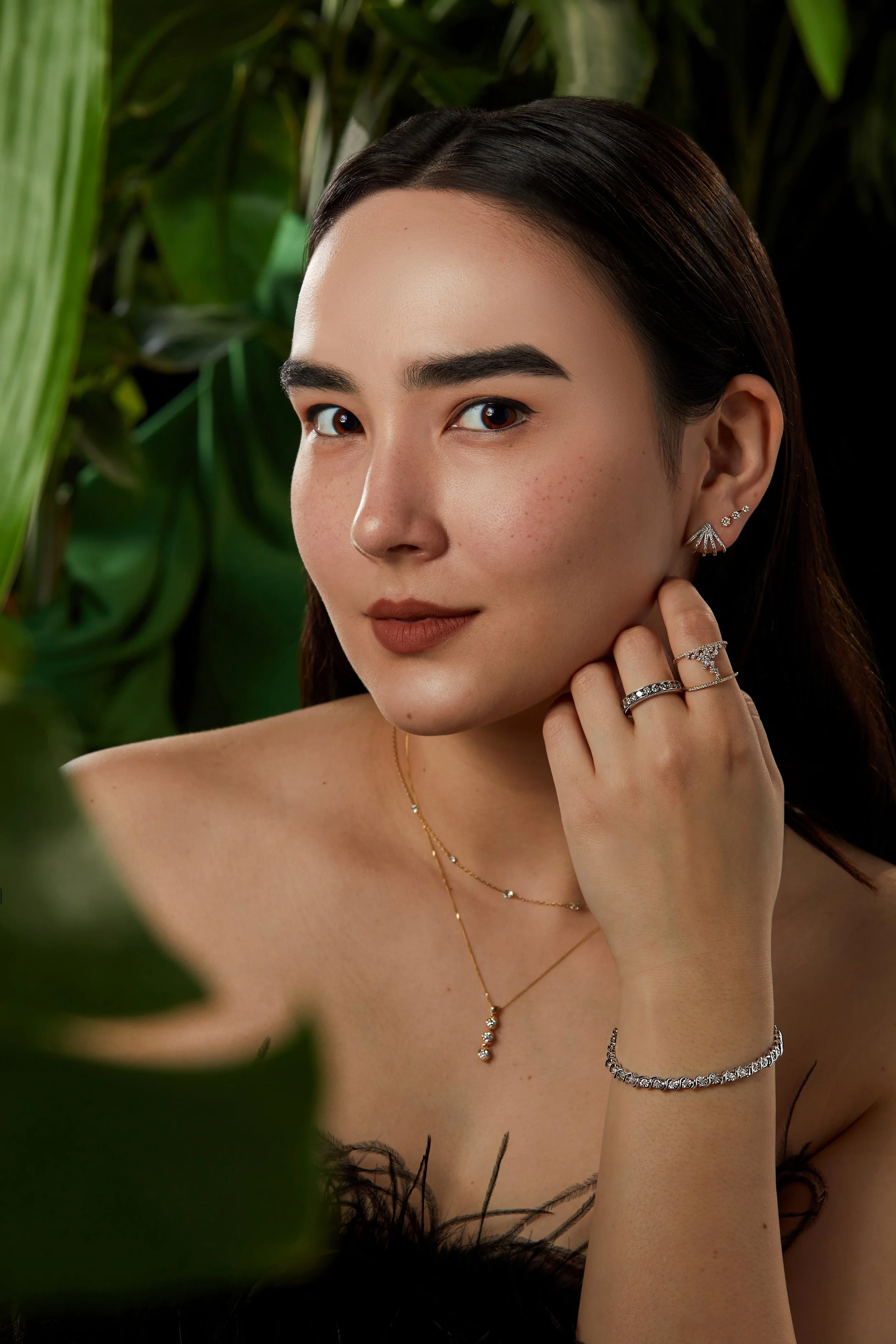 A young woman with long dark hair, wearing jewelry including earrings, rings, necklaces, and a bracelet, poses amidst green foliage, looking at the camera.
