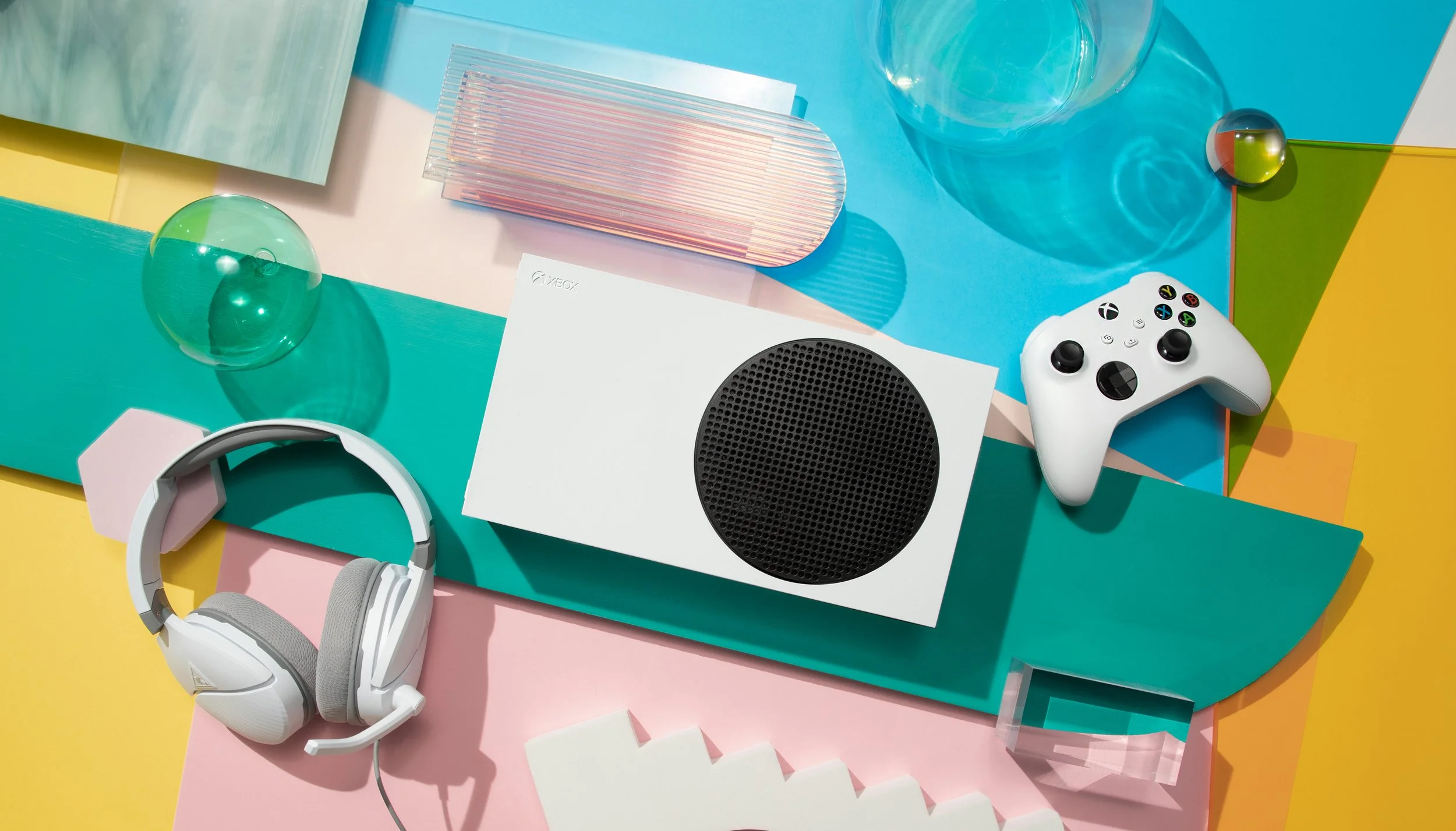 Flat lay of electronic gadgets and accessories, including a white gaming controller, white headphones, a black and white portable speaker, and transparent colorful acrylic sheets, arranged on pastel-colored surfaces with reflections and shadows.