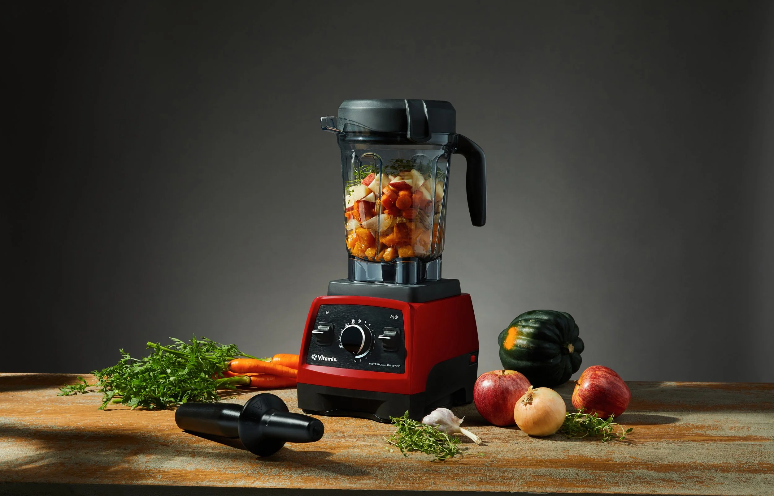 A red Vitamix blender filled with chopped vegetables on a wooden surface surrounded by fresh vegetables and herbs, including carrots, onions, garlic, and a green squash, with a dark gray background.