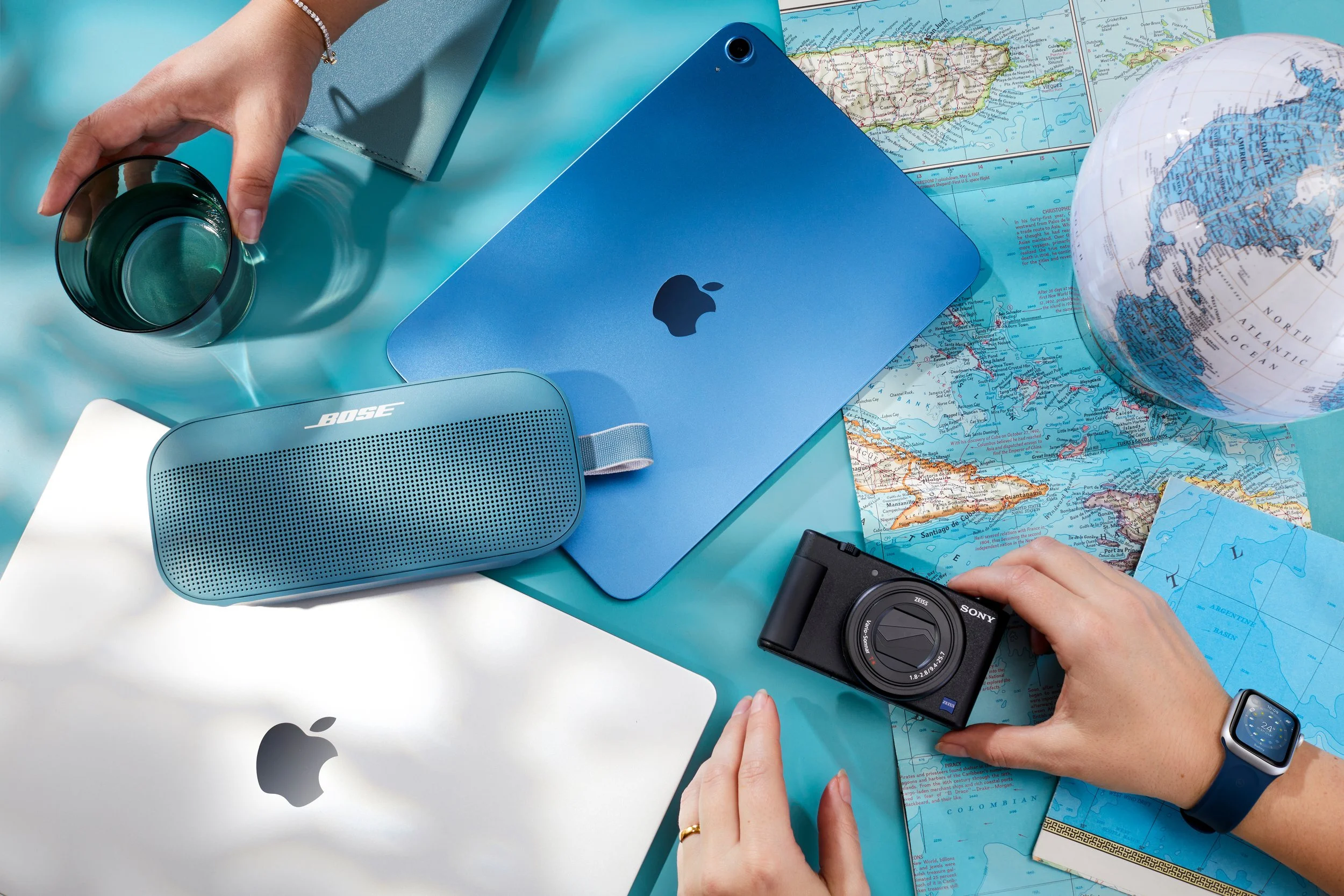 Travel essentials on a table, including maps, a globe, a laptop, a camera, a portable speaker, a glass of water, and a smartwatch.