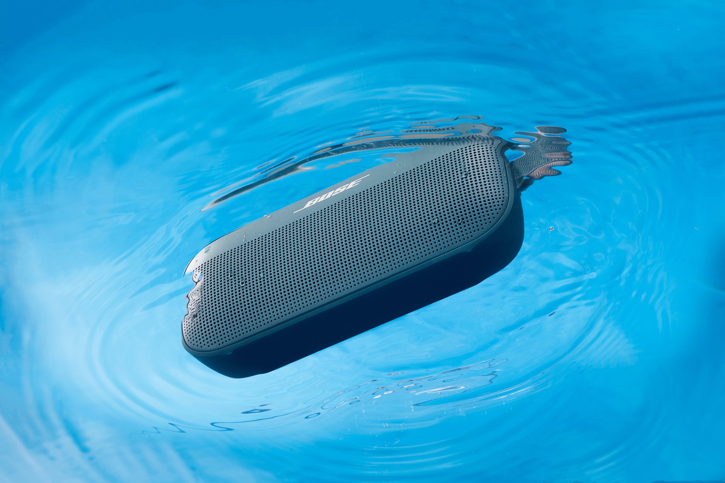 A black Bose portable speaker floating in a swimming pool, creating ripples on the water surface.