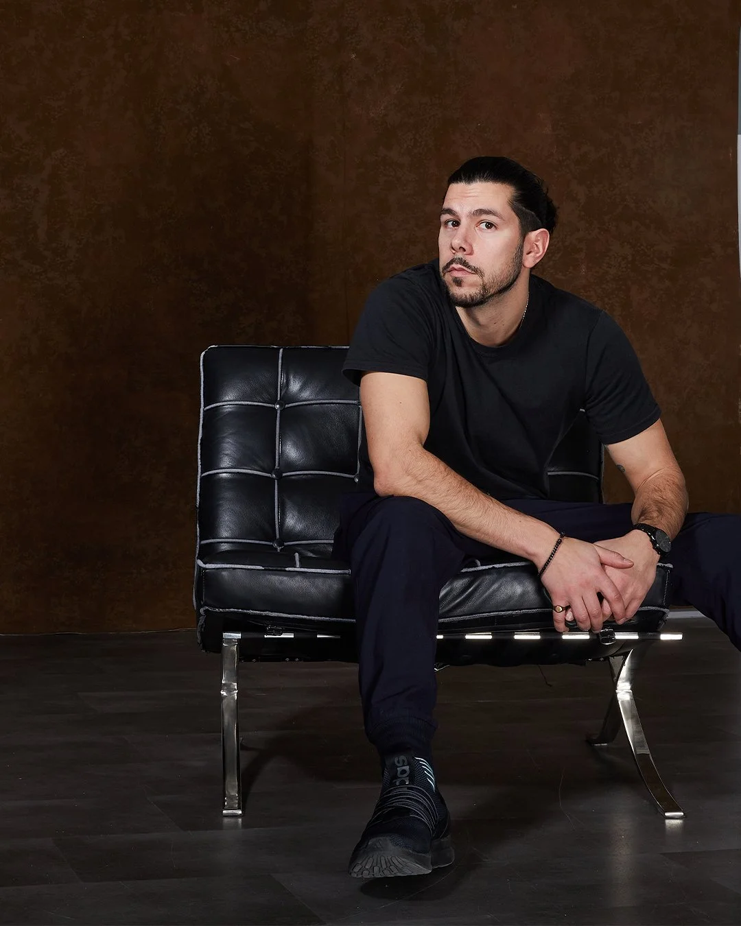 A man sitting on a modern black leather chair against a brown wall, wearing a black t-shirt, black pants, and black sneakers with a serious expression.
