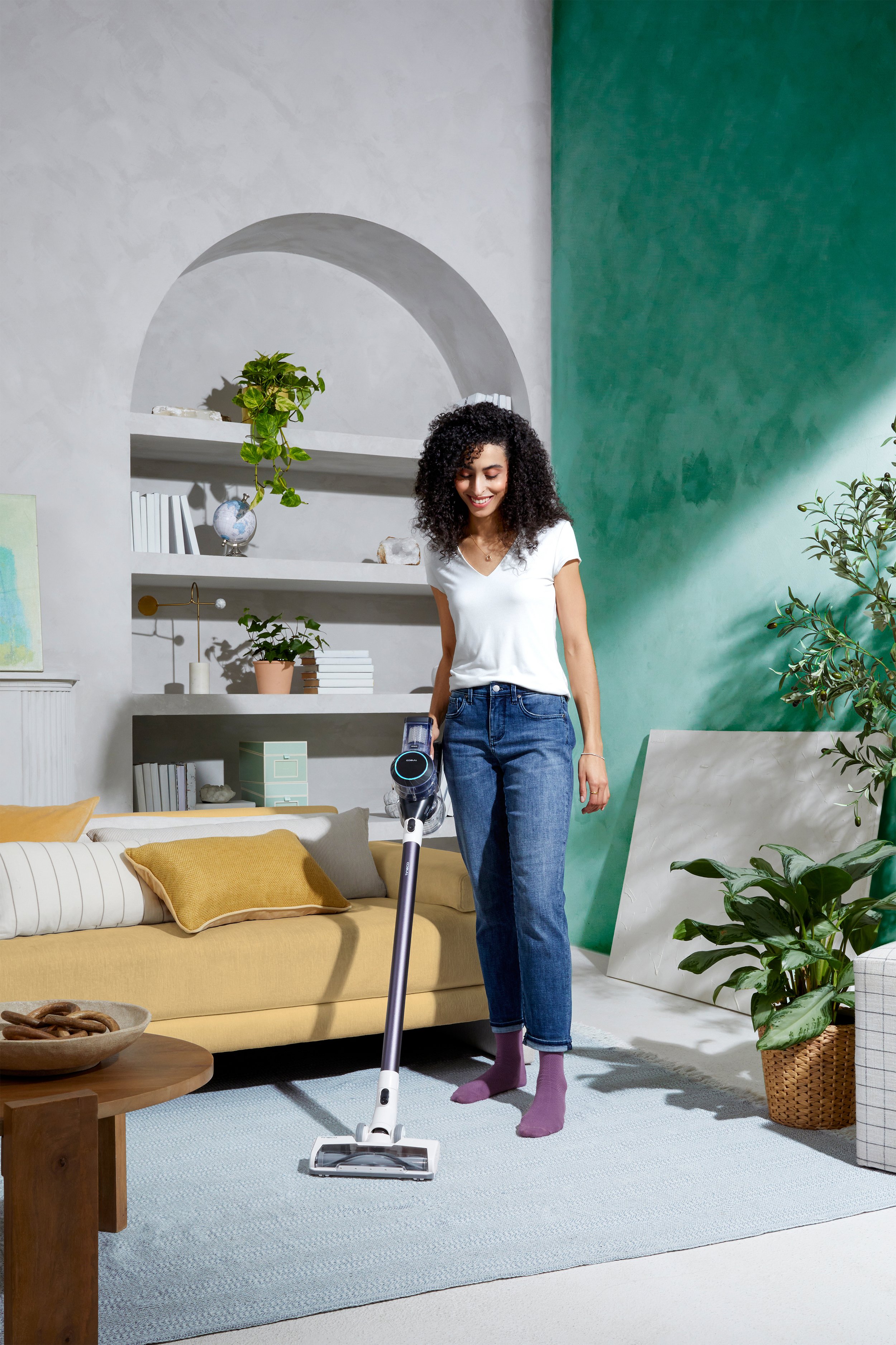 A woman vacuuming a living room with a stick vacuum cleaner. She is smiling and wearing a white t-shirt, jeans, and purple socks. The room has a yellow sofa, green plants, and white and green walls.