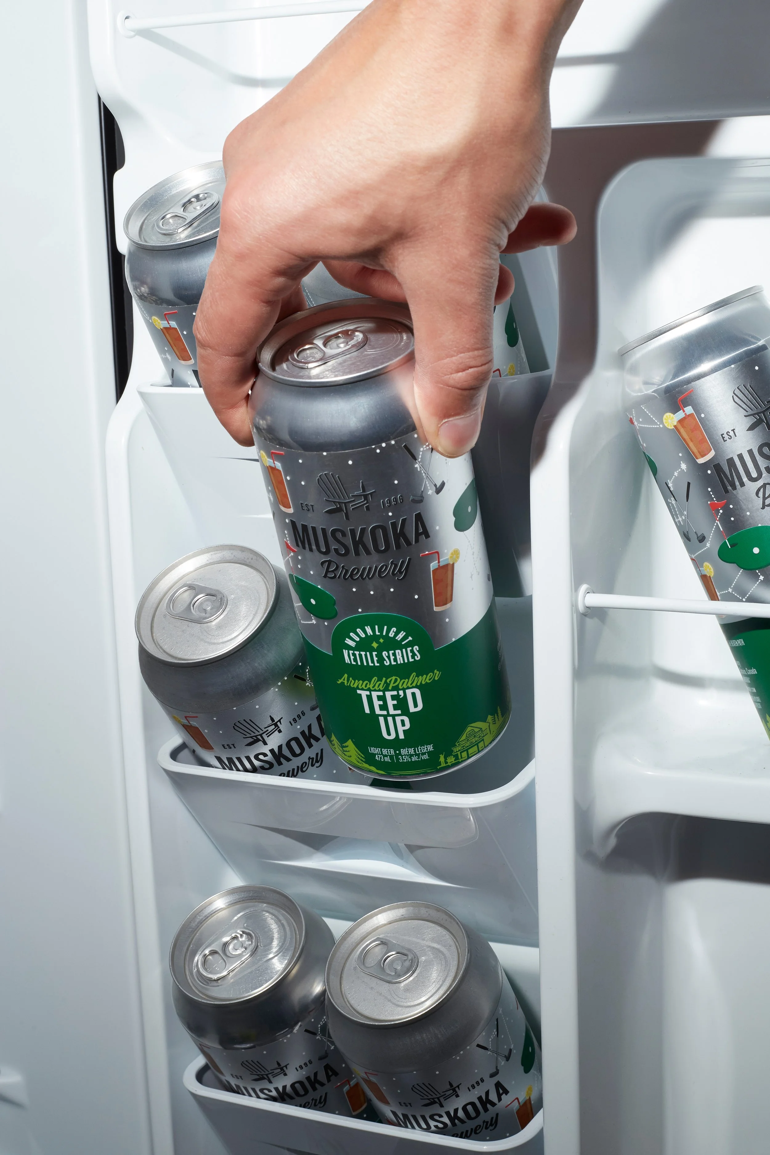 A hand placing a can of Muskoka Brewery beer labeled 'Tee'd Up' into a refrigerator.