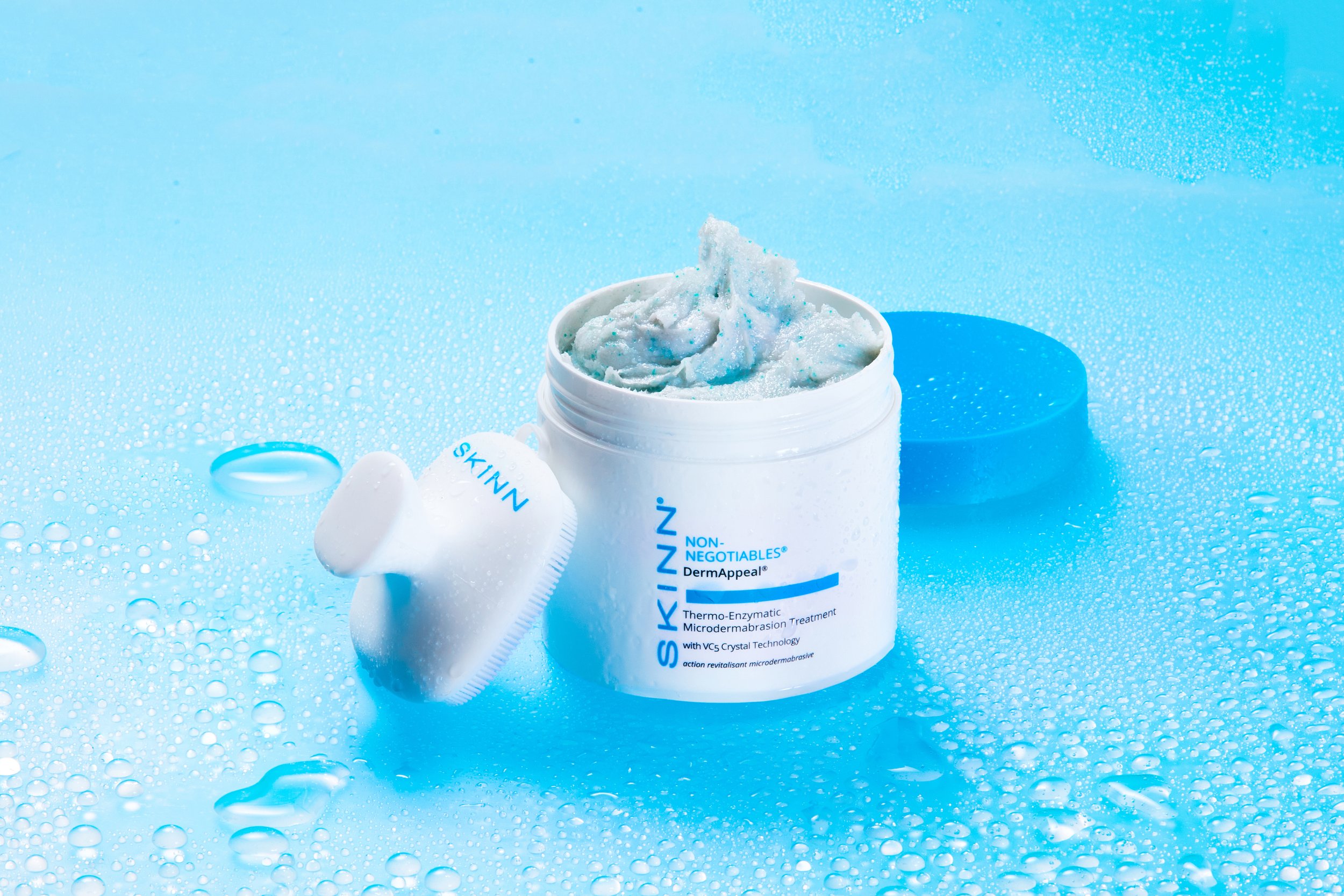 Skinn DermAppeal microdermabrasion treatment cream with a blue and white color scheme, open jar showing cream, a microdermabrasion brush applicator, and a blue lid on a wet blue surface with water droplets.