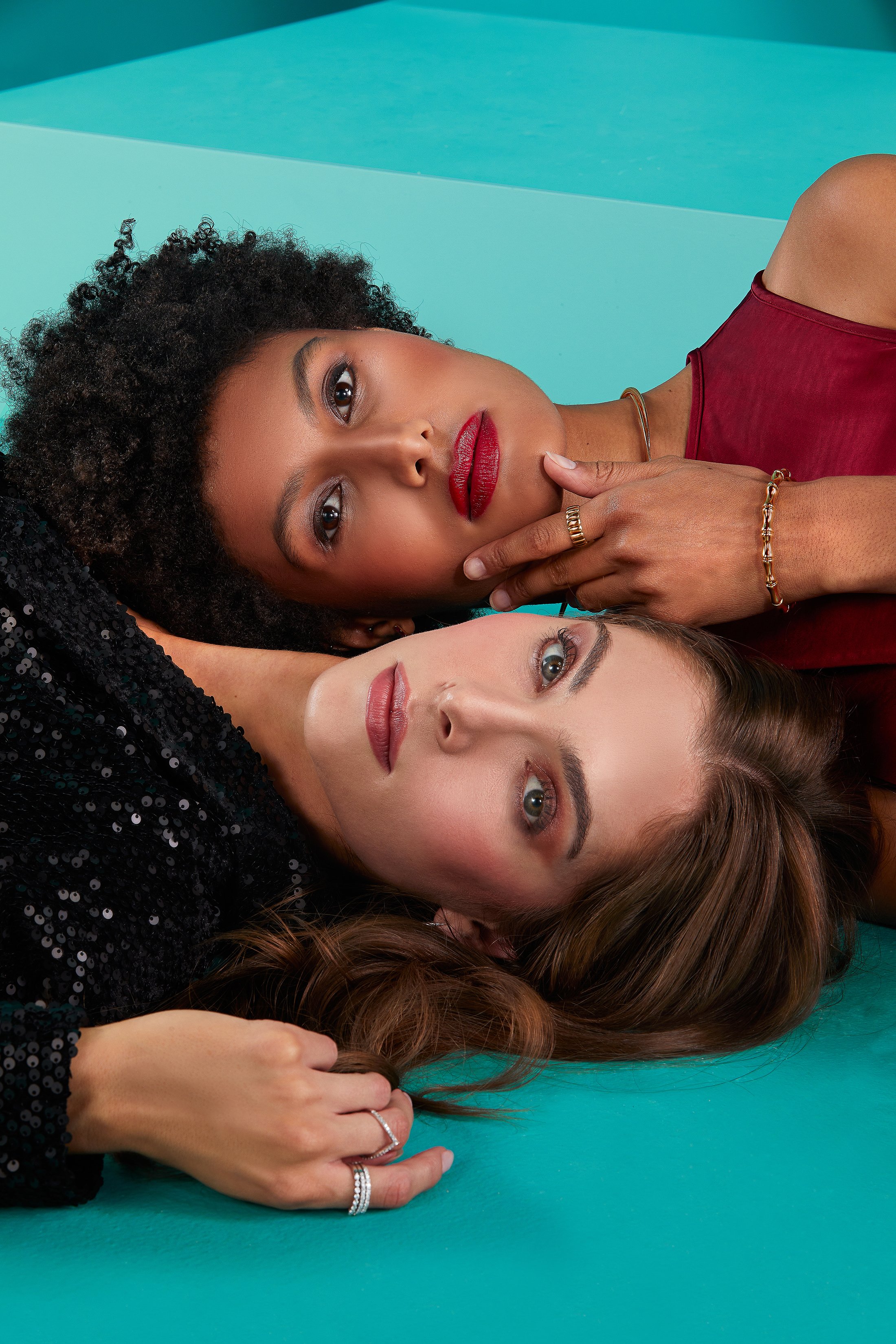 Two women lying on the floor, one with curly black hair and the other with straight brown hair, posing closely with serious expressions against a teal background.